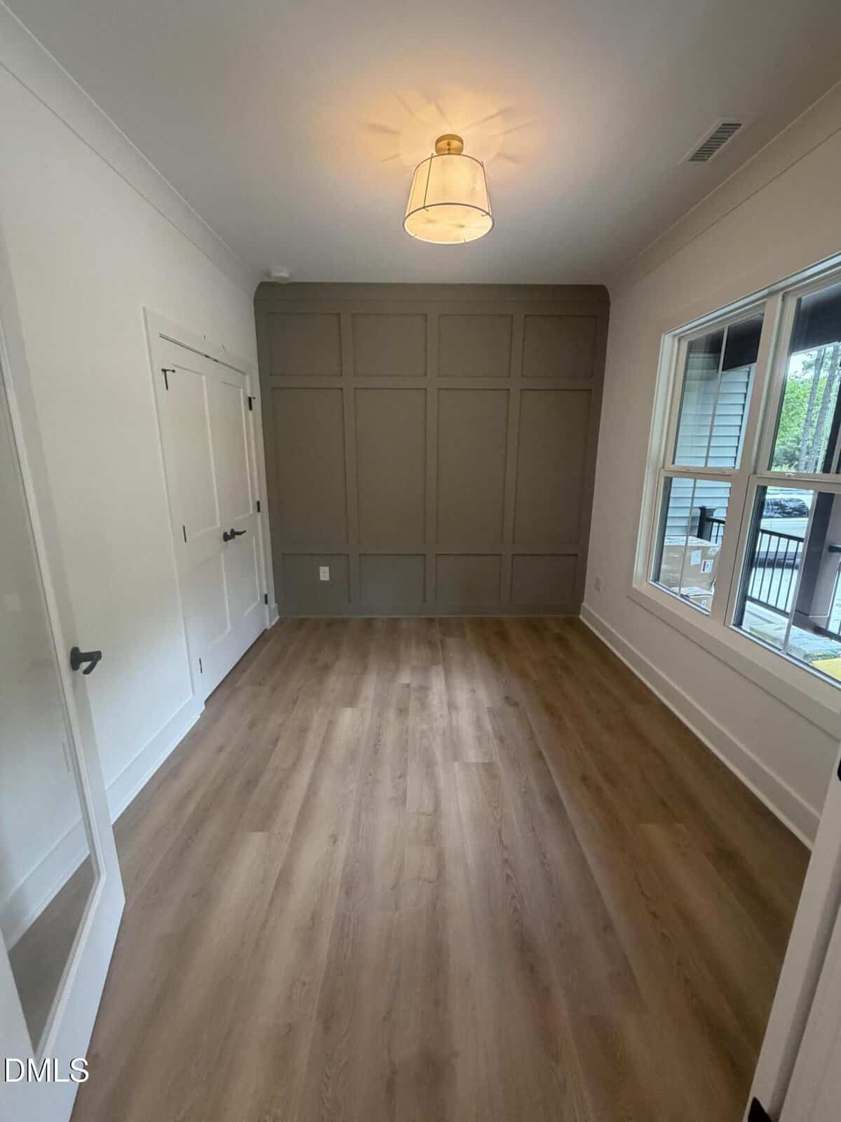 A small, empty room with light wood floors, white walls, a dark accent wall with grid paneling, a ceiling light fixture, double closet doors, and a window showing an outdoor scene.
