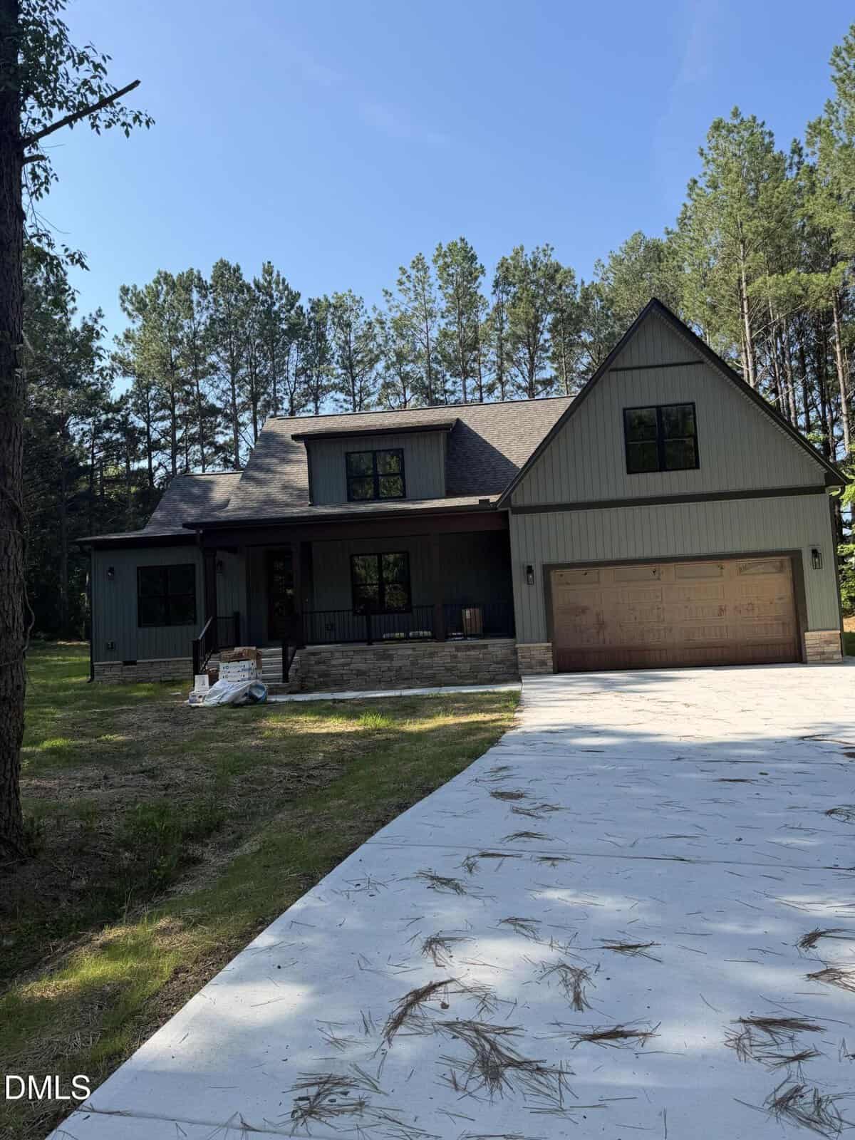A two-story house with a covered front porch and attached garage sits among tall pine trees. The driveway is concrete with scattered pine needles, and the sky is clear and blue.