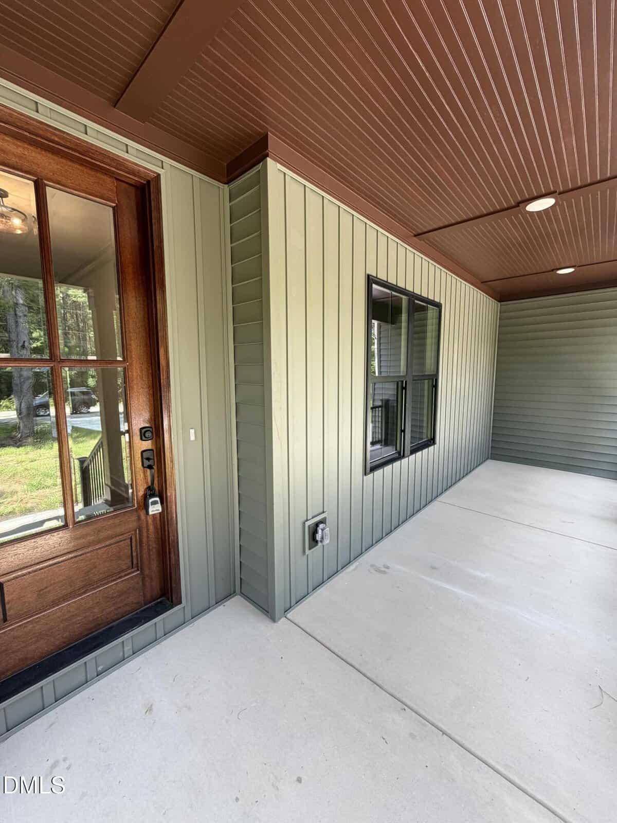 A covered porch with green vertical siding, a wooden door with glass panes, two black framed windows, and a concrete floor. The ceiling and trim are brown. An electrical outlet is mounted on the wall.