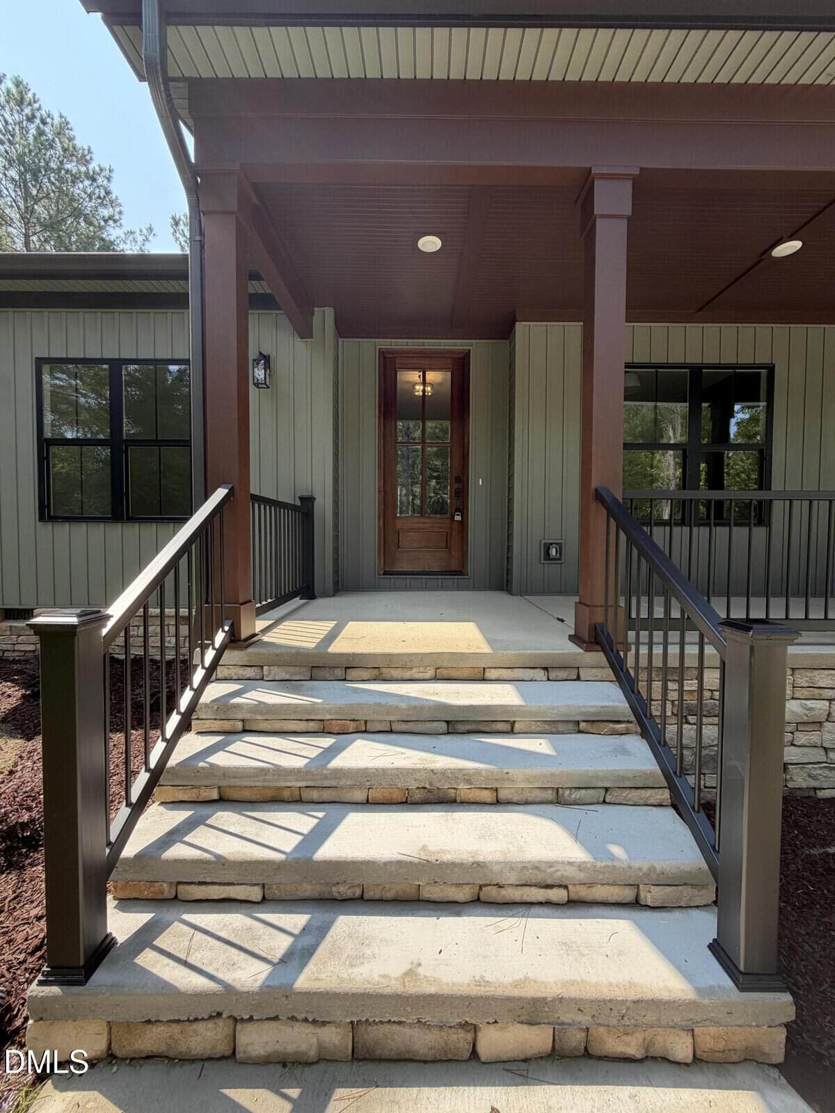 A front porch with stone steps leads to a wooden door. Metal railings border the steps, and the exterior features vertical siding with large windows. Trees are visible in the background.