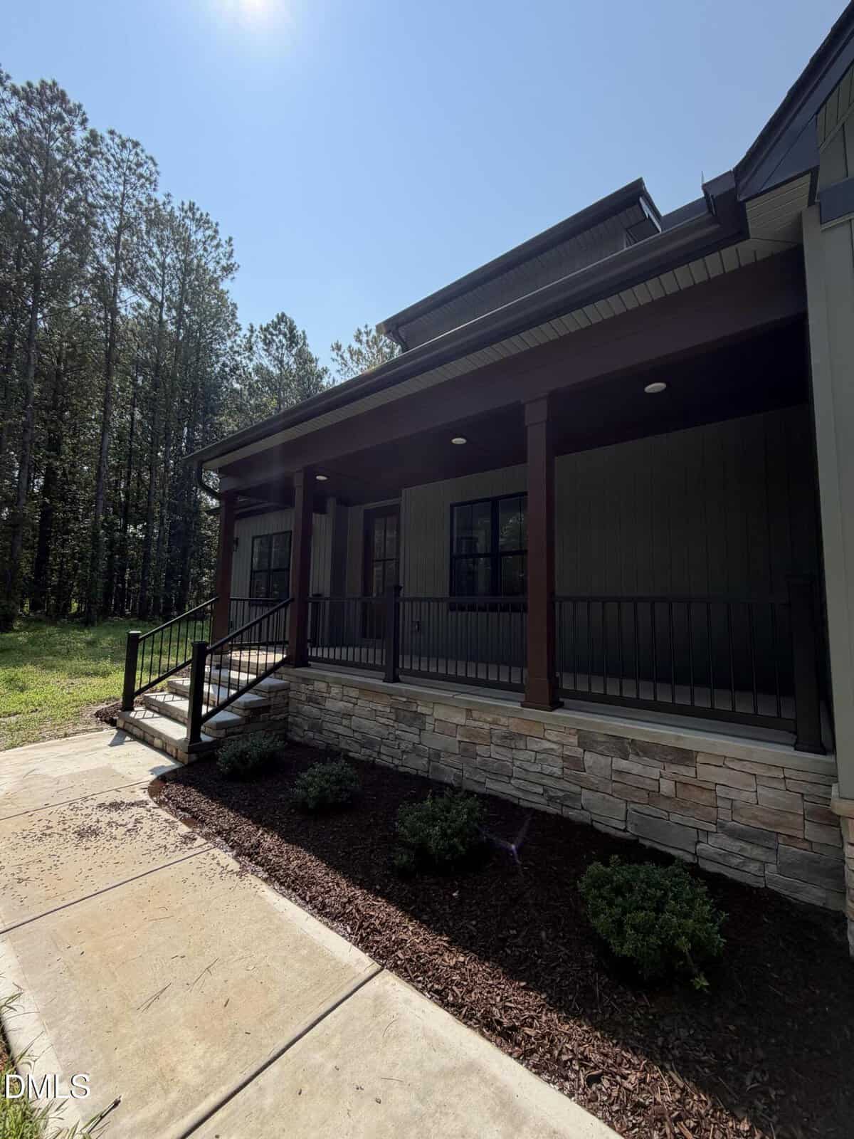 A house with a covered front porch, dark wooden columns, stone and mulch landscaping, black railing, and concrete walkway. Trees and grass are visible in the background under a sunny sky.