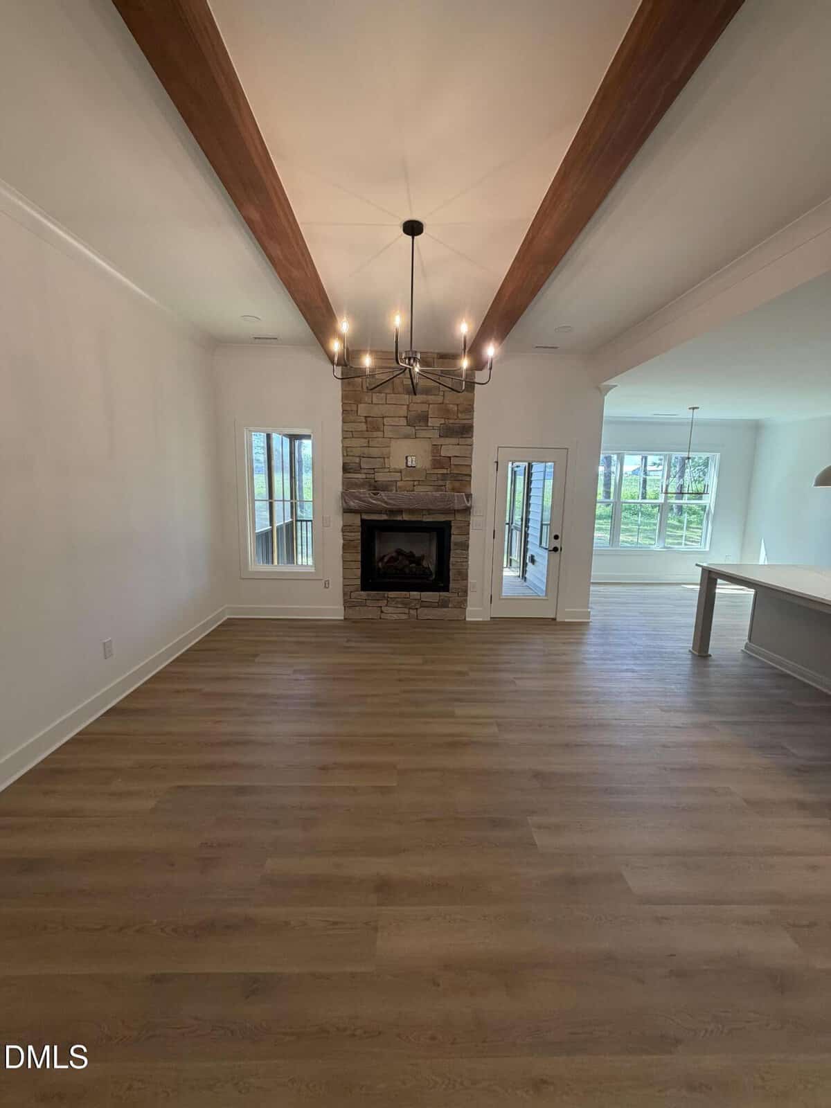 A spacious, empty living room with light wood floors, a stone fireplace, a black chandelier, wood ceiling beams, and large windows letting in natural light.