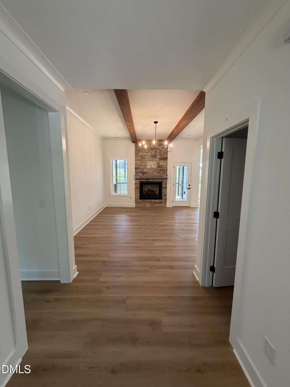 A hallway with light wood floors leads to a living room featuring a stone fireplace, exposed wood ceiling beams, two windows, and a modern chandelier. White walls and doors frame the space.