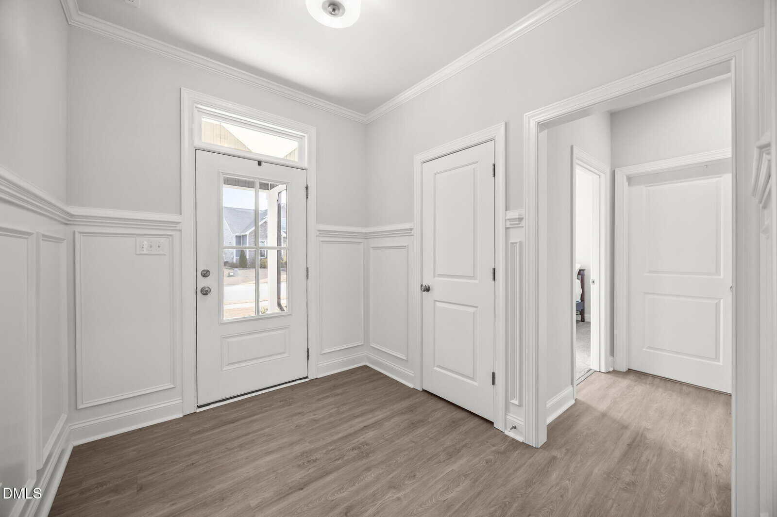 A bright, empty entryway with white walls, wainscoting, light wood flooring, a glass-paneled front door, and three interior doorways leading to other rooms. Natural light enters through the front door window.