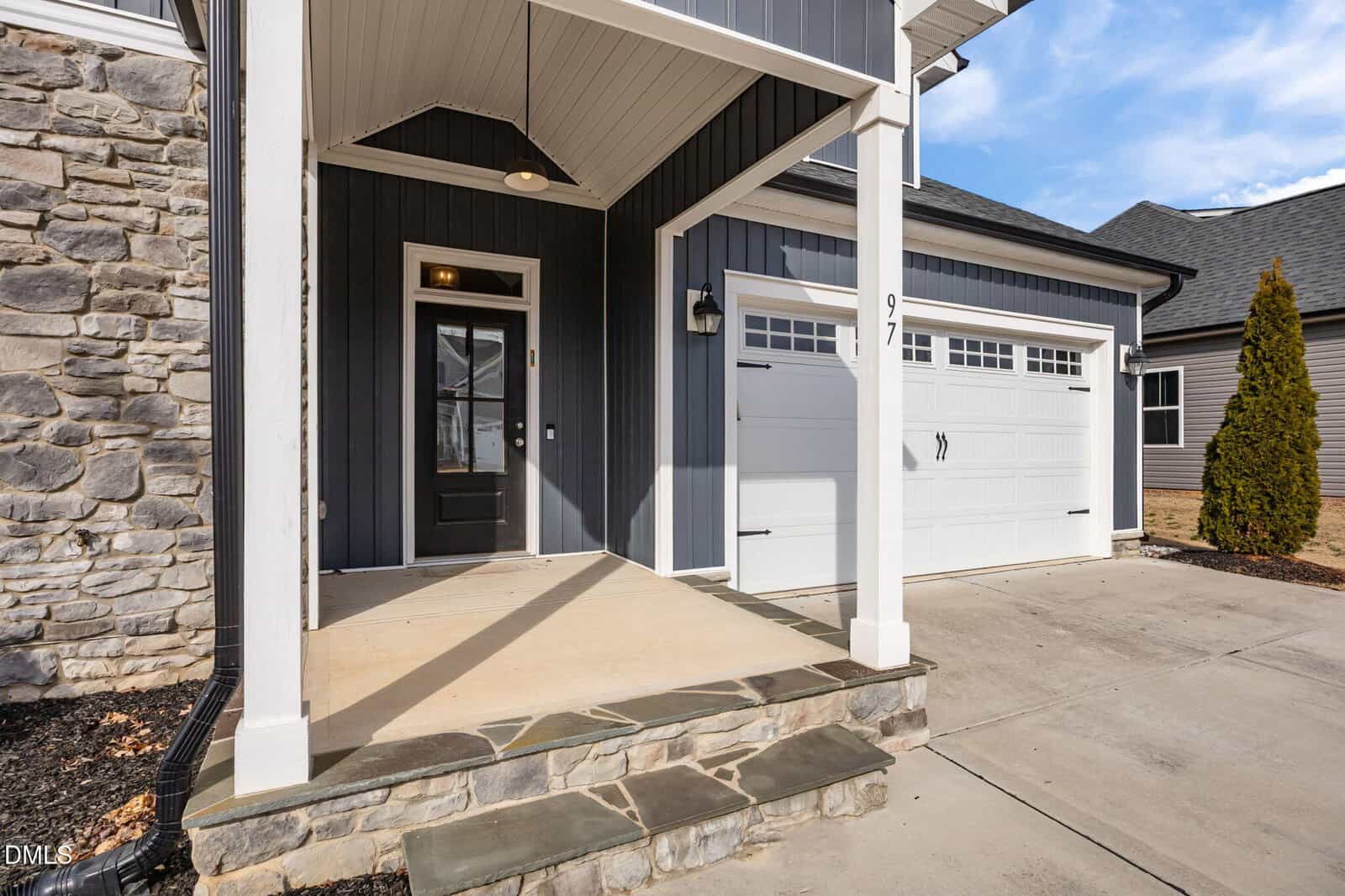 Front entrance of a modern house with dark siding, stone accents, a covered porch, white garage door, and concrete driveway; the porch has steps and a black front door with sidelights.