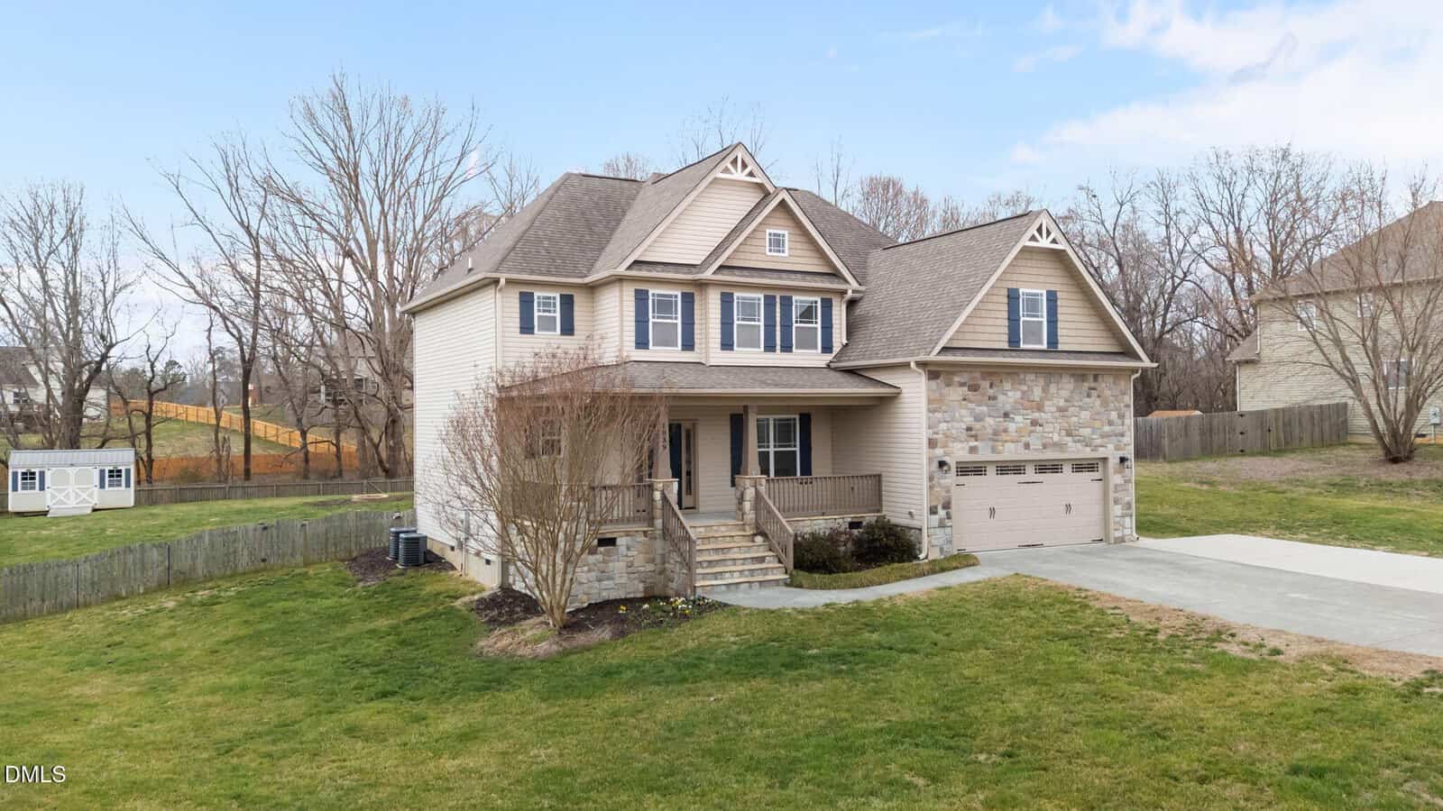Two-story suburban house with beige siding, stone accents, and a two-car garage. The property features a covered front porch, concrete driveway, sparse landscaping, and a spacious, grassy yard with leafless trees and a fenced area in the background.