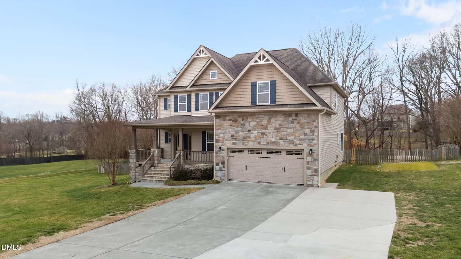 Two-story suburban house with stone and siding exterior, covered front porch, double garage, and a wide driveway. The yard has grass and trees, with a wooden fence enclosing part of the backyard. The sky is clear.