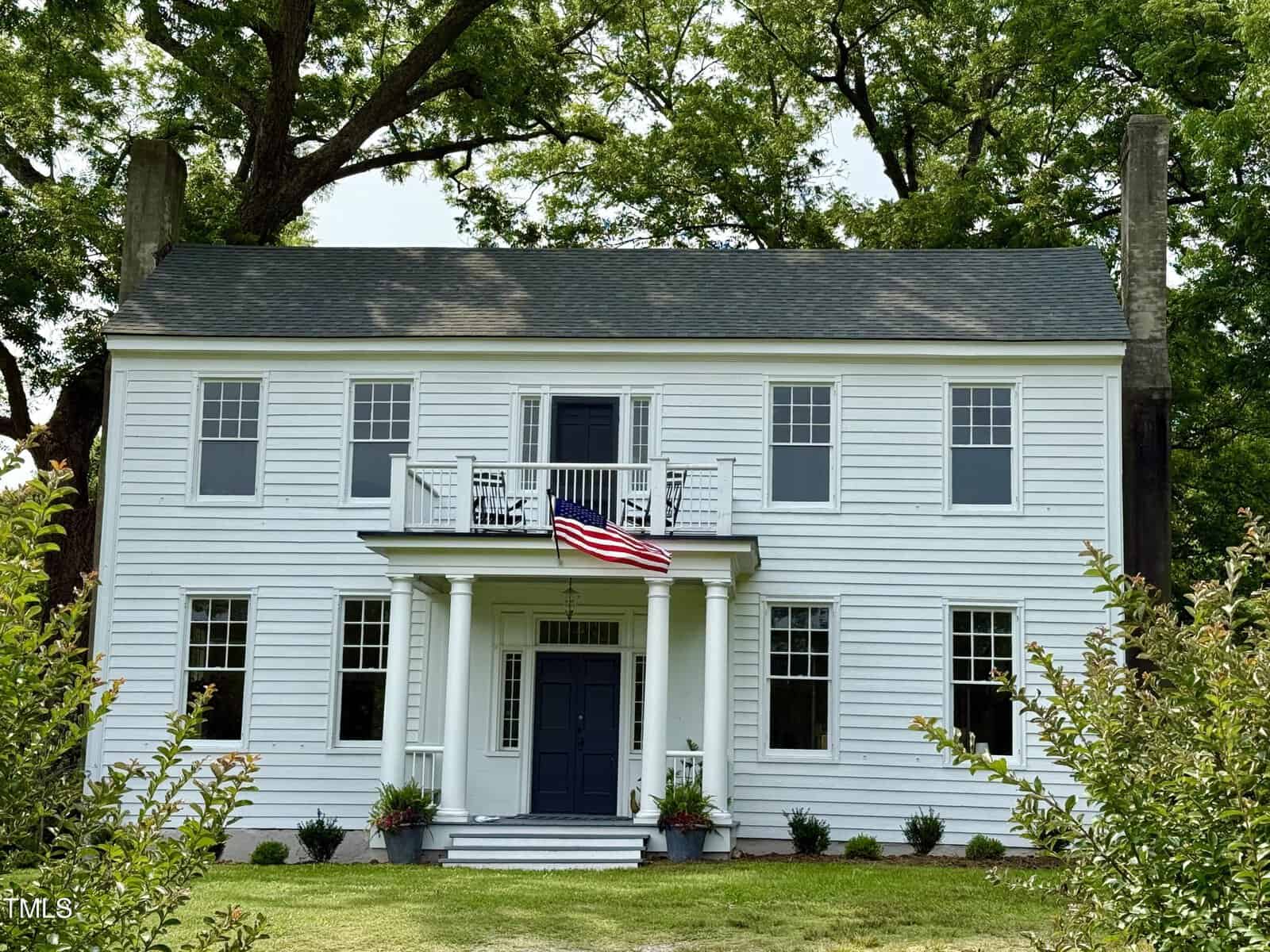A two-story white colonial-style house with black shutters, a central dark door, columns at the entrance, an American flag above the porch, and green trees and bushes surrounding the building sits gracefully at 313 W Noble Street in Louisburg.