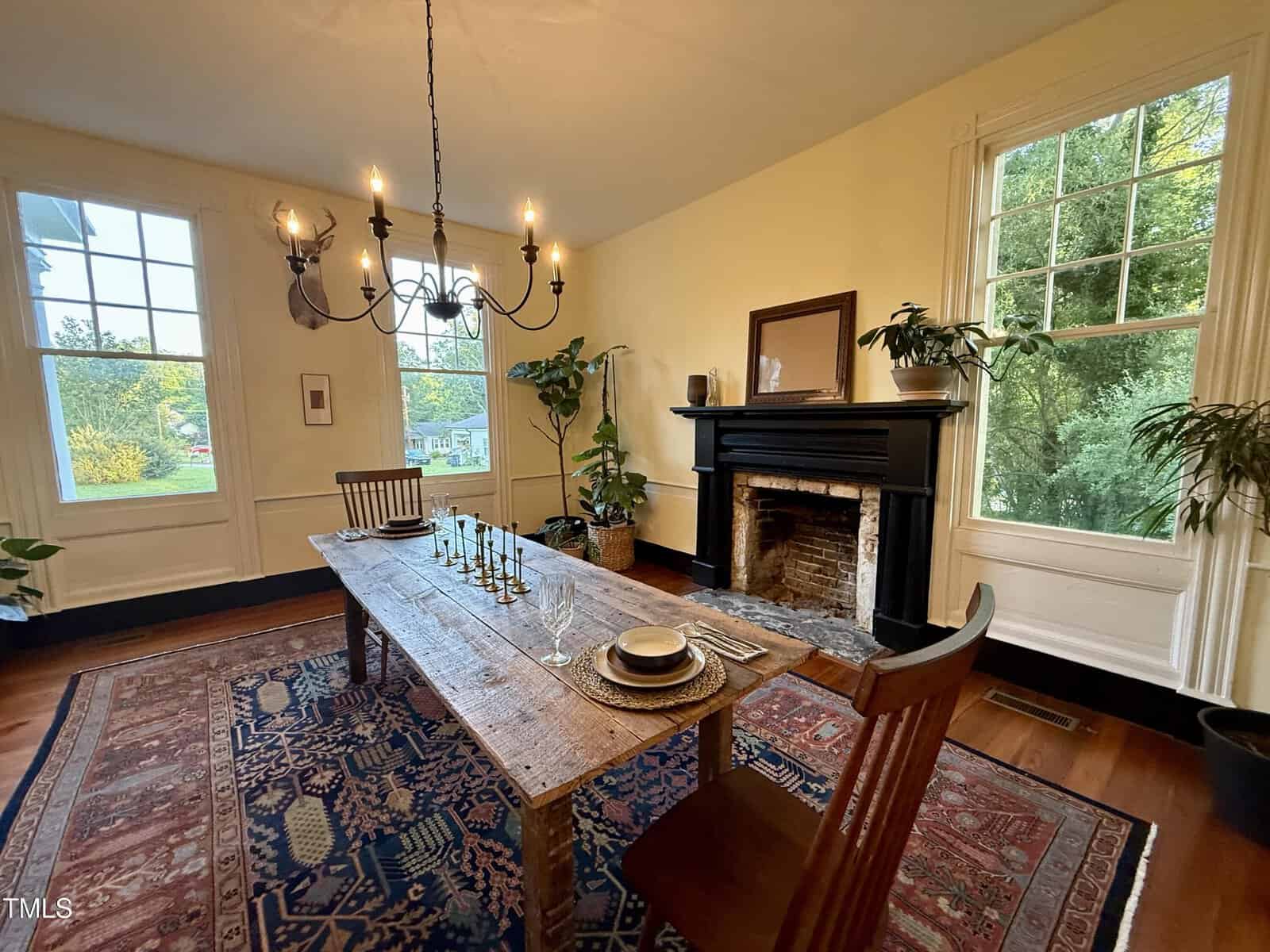 A dining room with a rustic wooden table, two chairs, a fireplace, houseplants, large windows letting in natural light, and a patterned rug on hardwood floors.