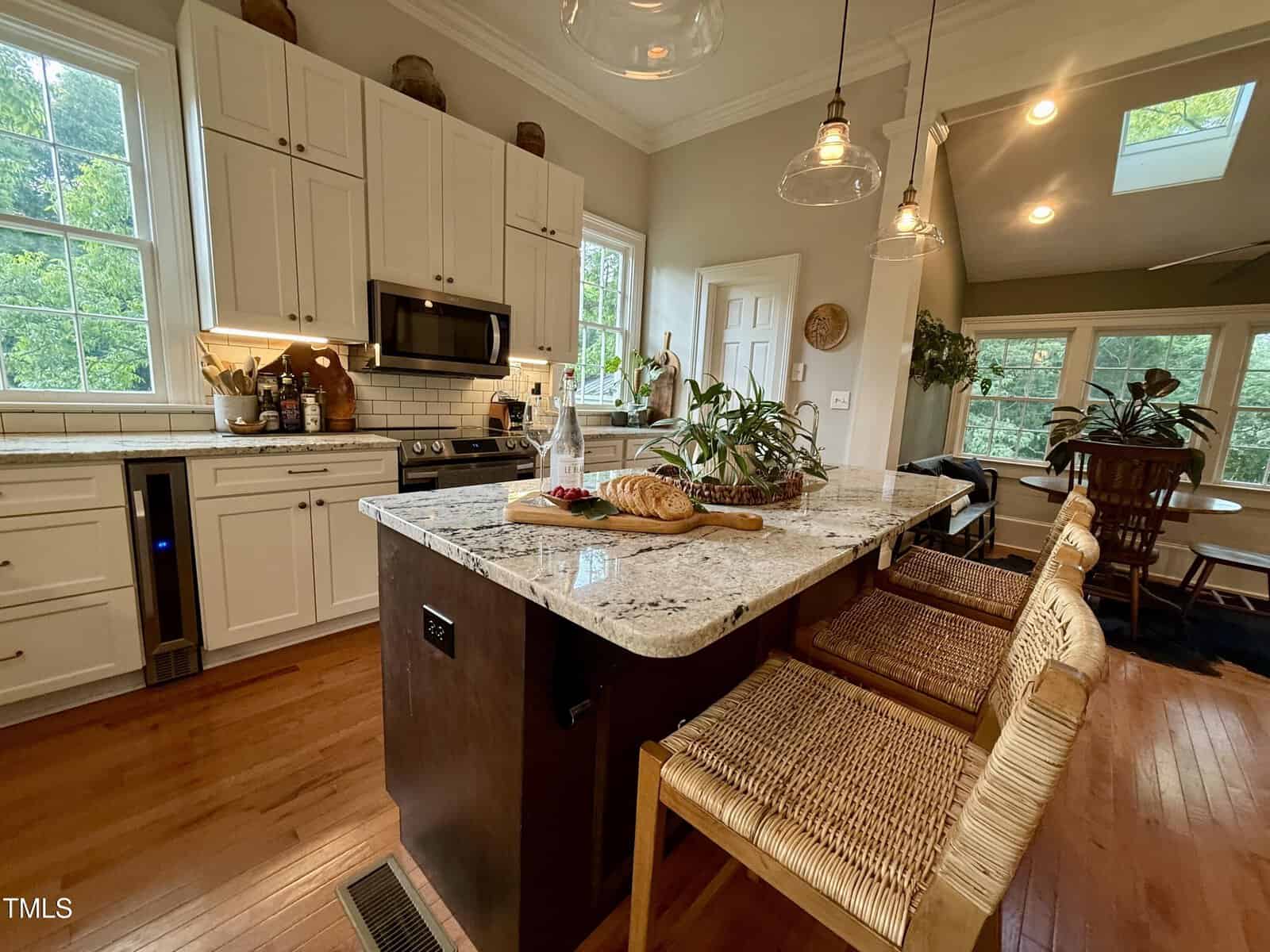 Modern kitchen with white cabinets, stainless steel appliances, and a large island with granite countertop. Two wicker barstools, bread, and plants are on the island. Sunlit windows and a dining area are visible in the background.