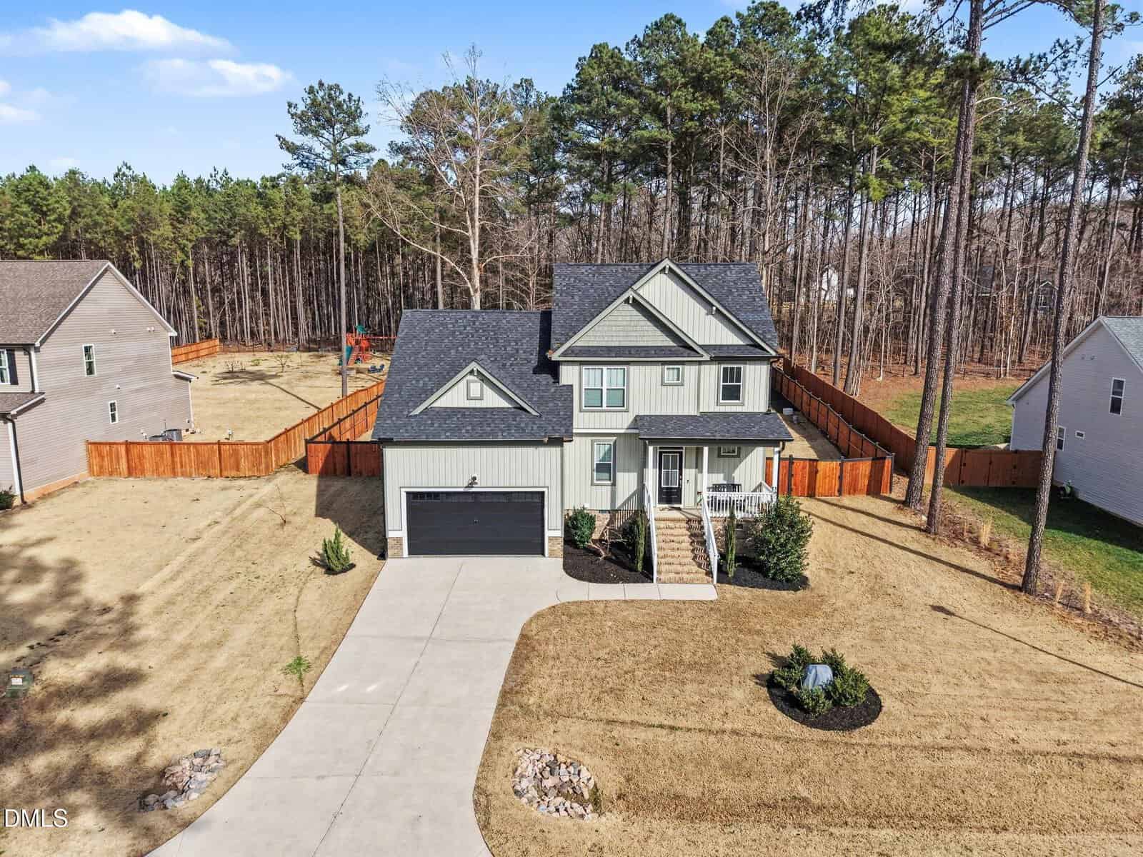 Two-story house with a gray exterior, black roof, attached two-car garage, front porch, and fenced backyard, situated on a large lot with pine trees in the background and neighboring homes on each side.