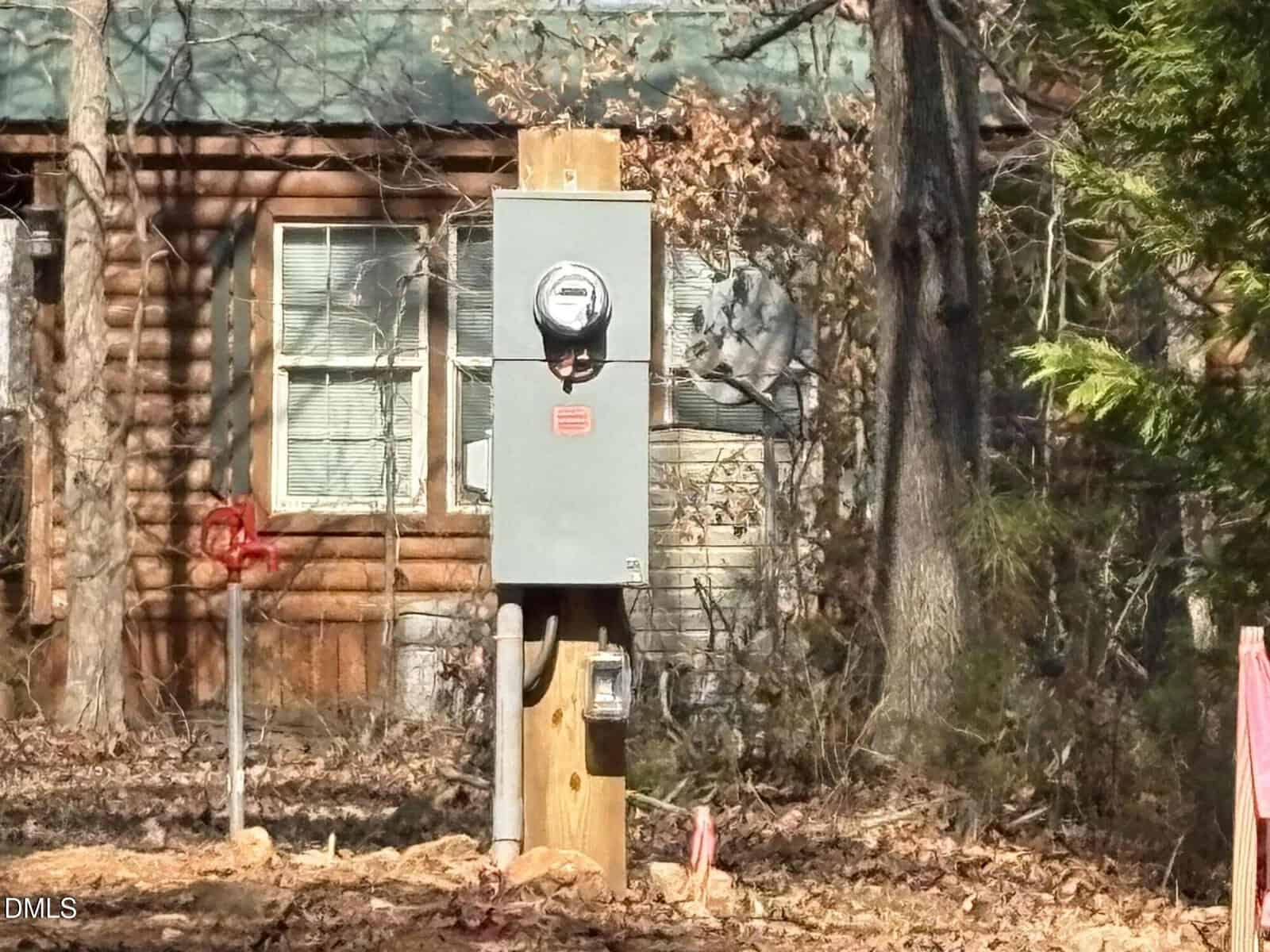 A freestanding electric meter and utility box are installed on a wooden post in front of a log cabin with windows, surrounded by trees and dry leaves.