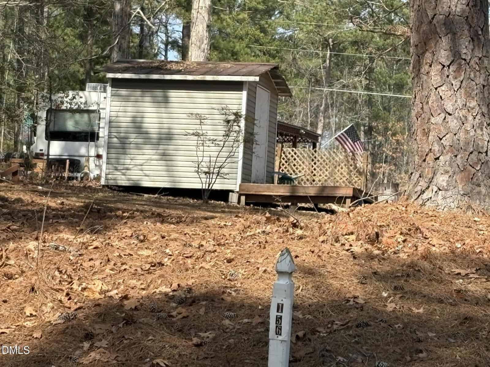 A small shed with a porch sits among pine trees; a lattice fence and American flag are visible. An RV is partially seen behind the shed, and a white utility marker stands in the foreground among pine needles.