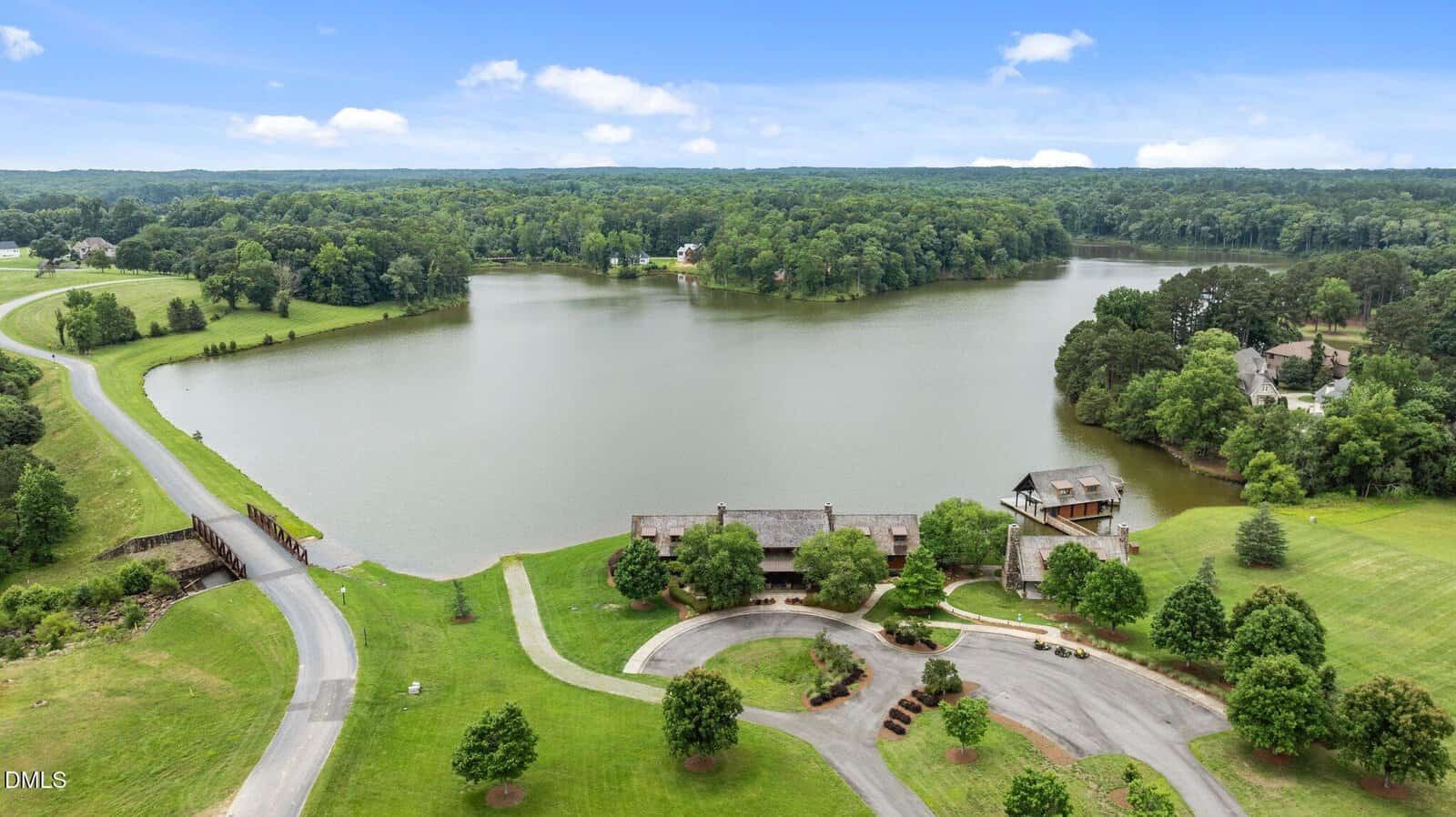 Aerial view of a scenic lakeside area with green lawns, trees, a curved road, and several large buildings near the water, surrounded by dense forests under a partly cloudy sky.
