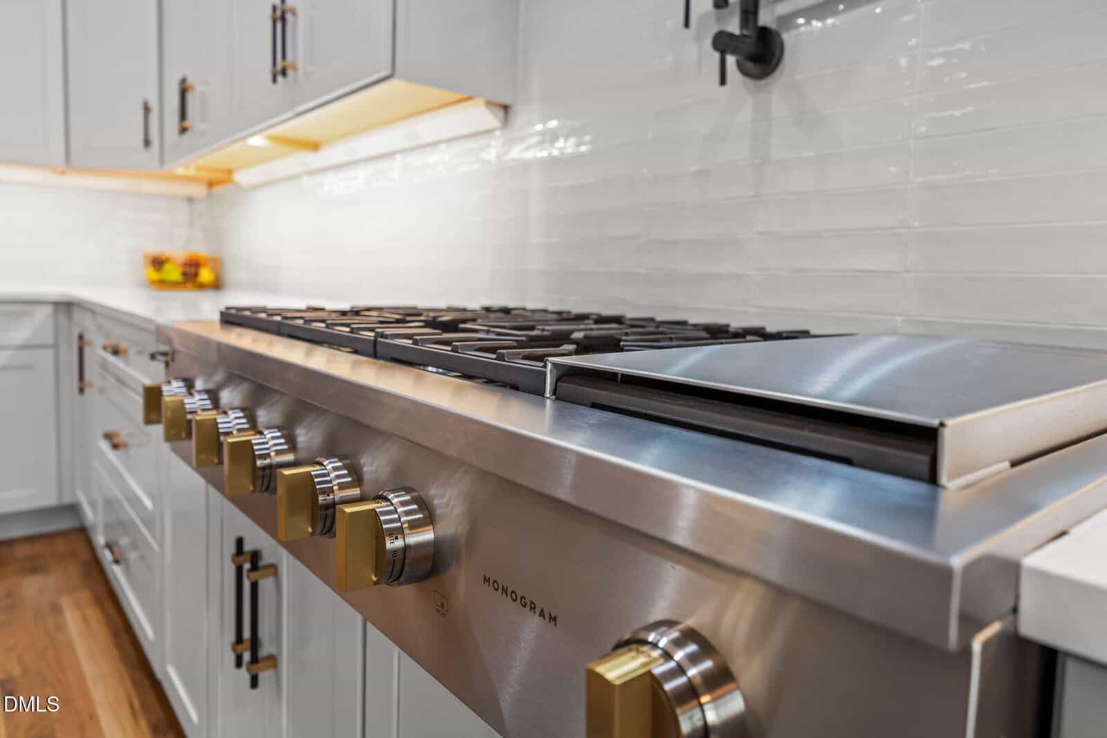 Close-up of a stainless steel Monogram gas stove with gold-colored knobs in a modern kitchen with white cabinets and a white tile backsplash.