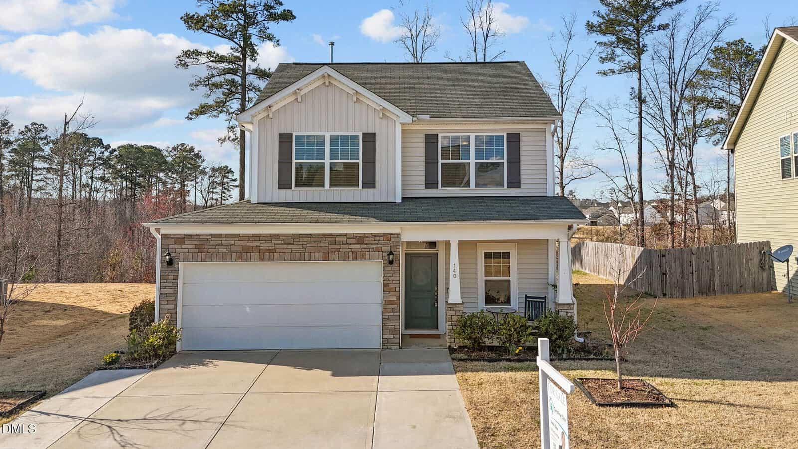 Two-story suburban house with white siding, stone accents, and an attached two-car garage. A covered front porch and small landscaped yard are visible, with trees and a wooden fence in the background.