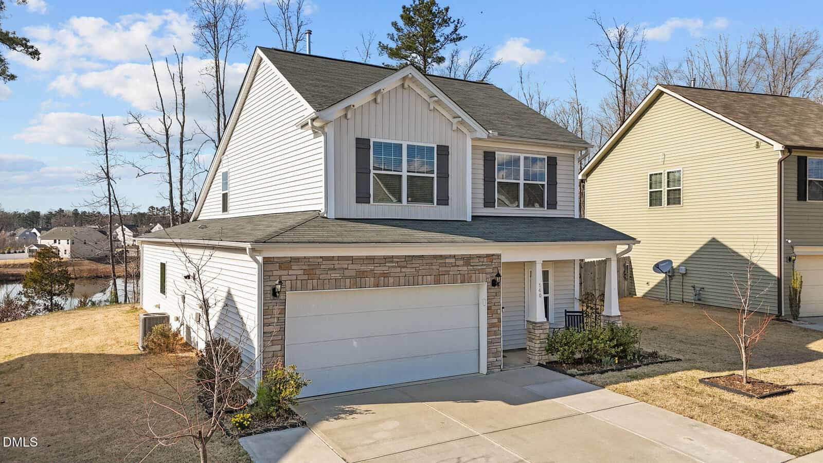 Two-story suburban house with gray siding, stone accents, and a double garage. The home has a small front porch and neatly trimmed lawn, with neighboring houses and a pond visible in the background under a blue sky.