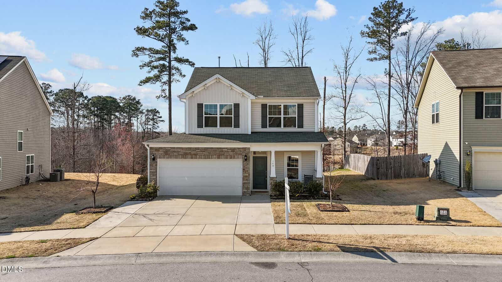 A two-story suburban house with a single-car garage, white siding, and brick accents. The house sits between two similar homes, with a paved driveway, manicured lawn, and trees in the background under a blue sky.