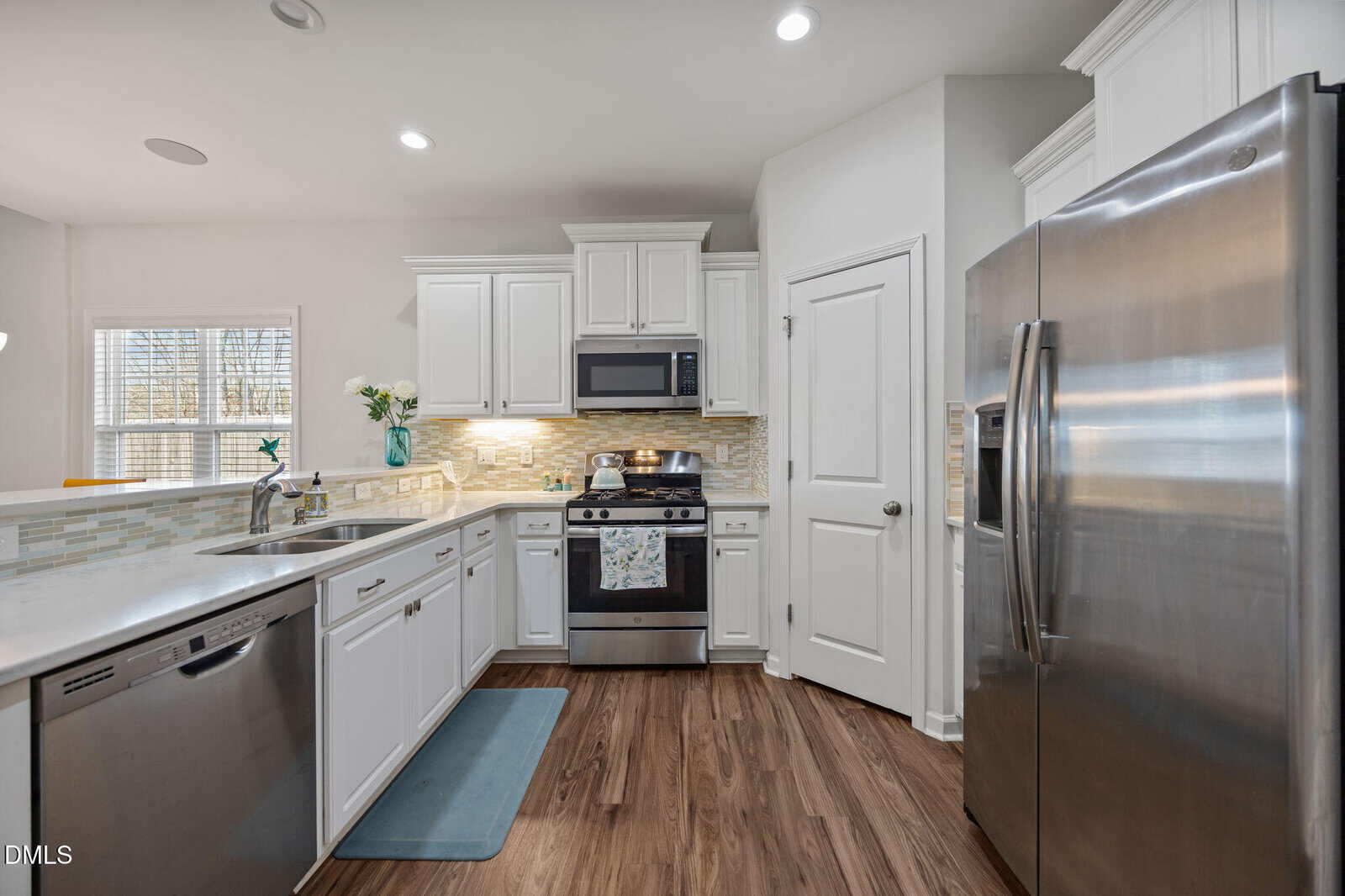 Modern kitchen with white cabinets, stainless steel appliances, wood flooring, tiled backsplash, sink, stove, refrigerator, and a window letting in natural light. A blue mat is placed by the sink.