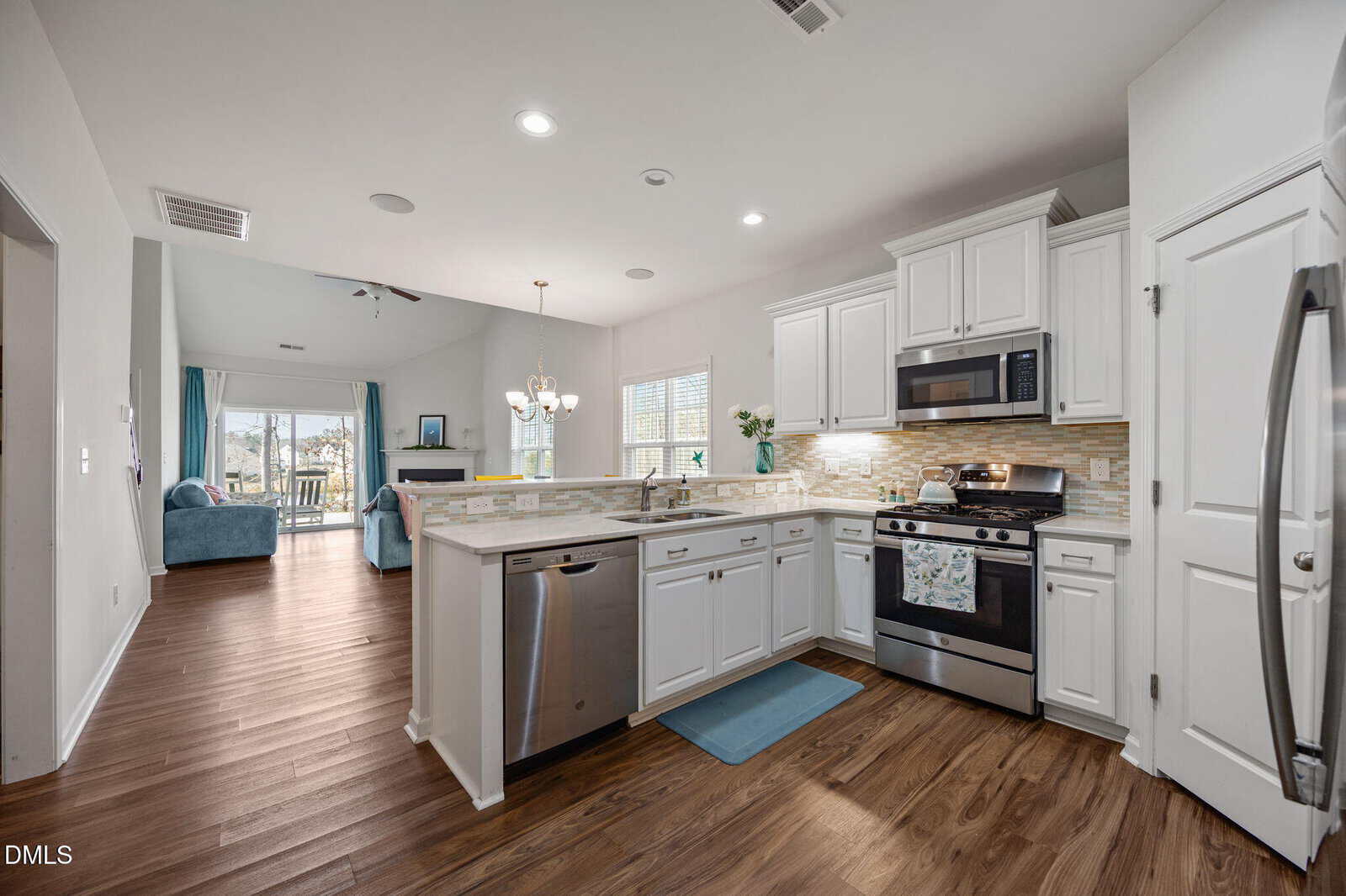 Modern kitchen with white cabinets, stainless steel appliances, and wood flooring, opening to a bright living and dining area with large windows and blue accents.