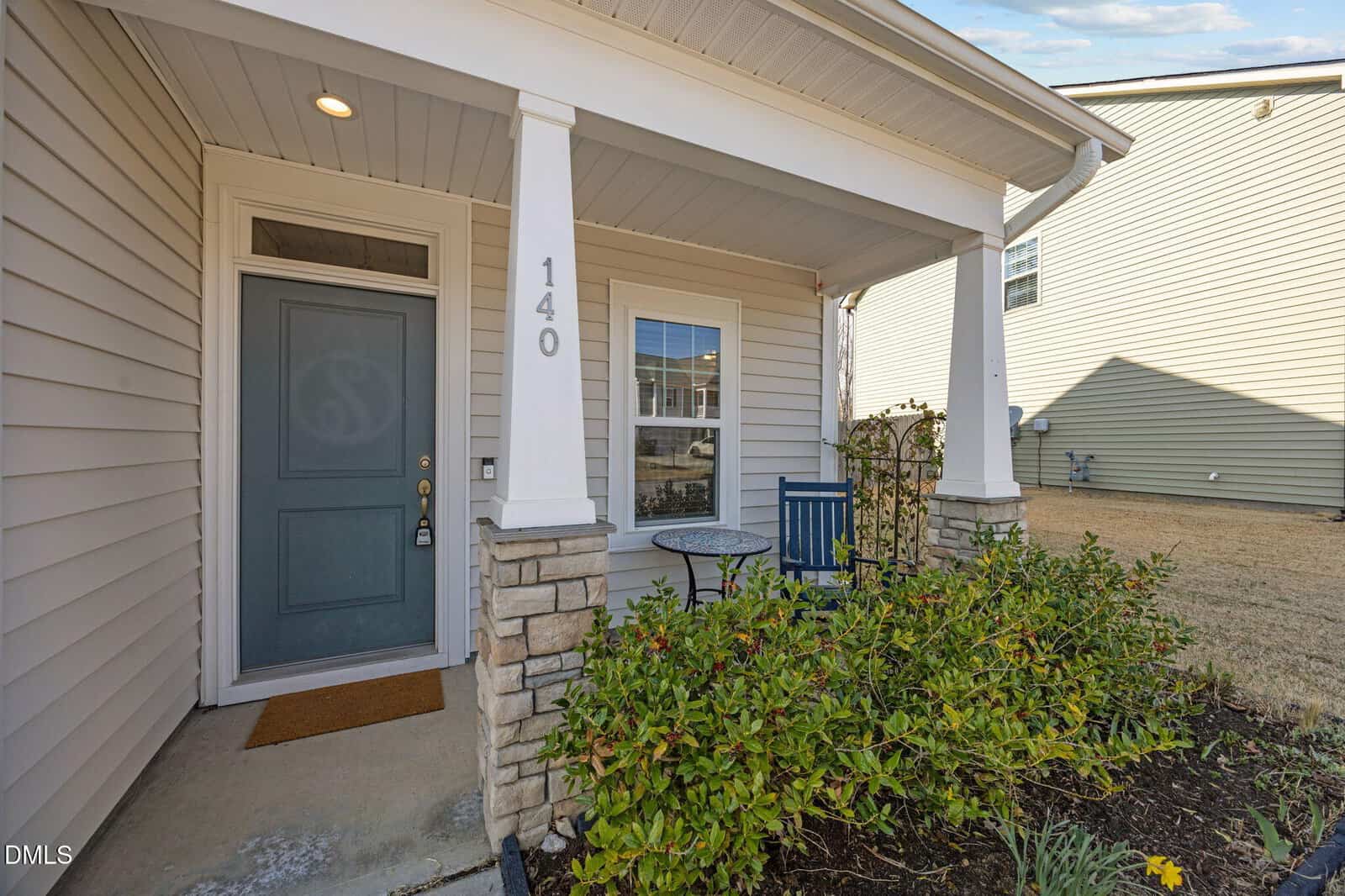 A small covered front porch with white columns, a blue door labeled "140," a window, and a blue bistro table with two chairs. Green shrubs and a welcome mat are by the entrance.