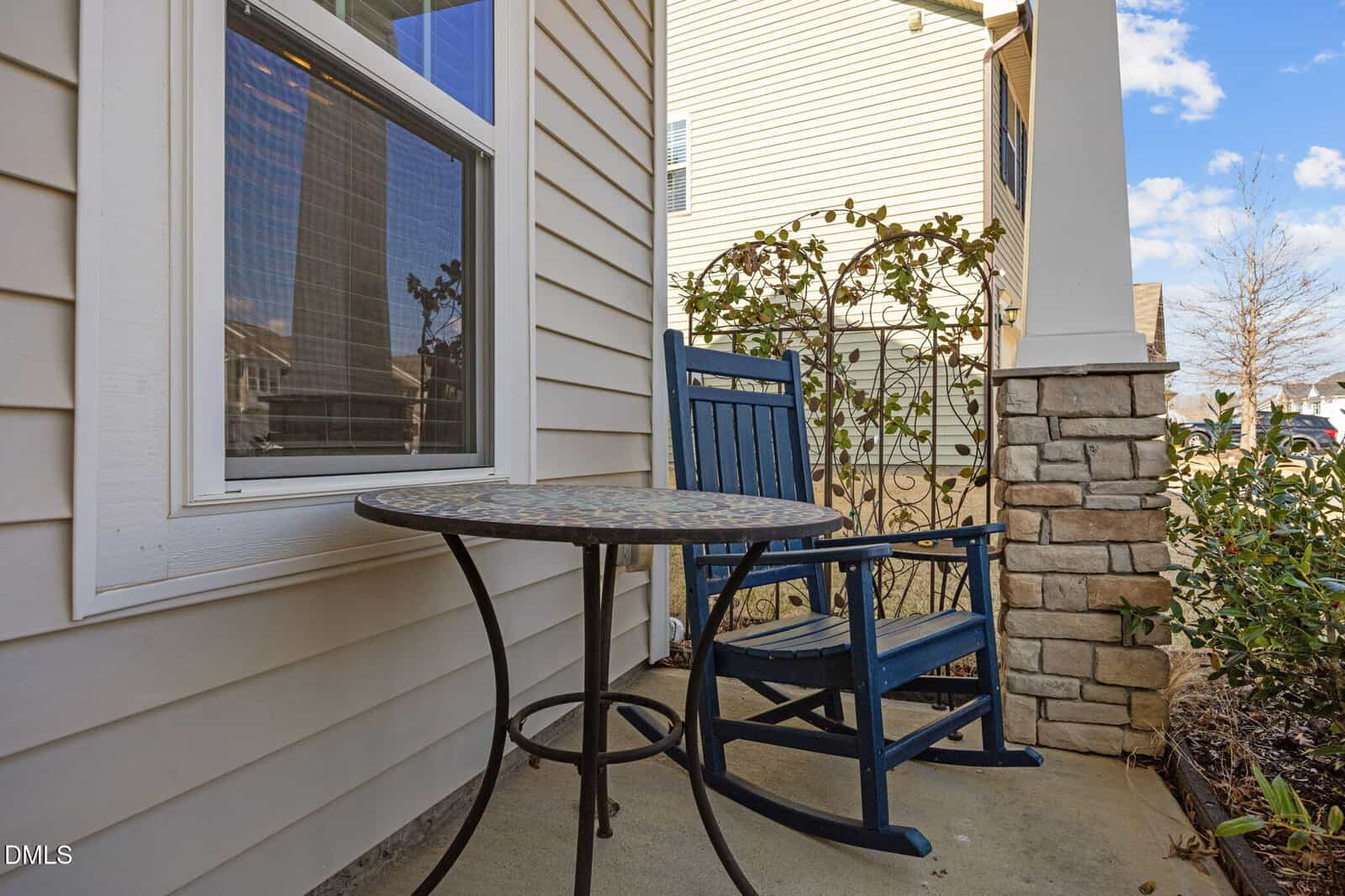 A small patio with a blue rocking chair and round metal table, next to a house with beige siding and a window. A stone pillar and lattice with climbing plants are nearby.