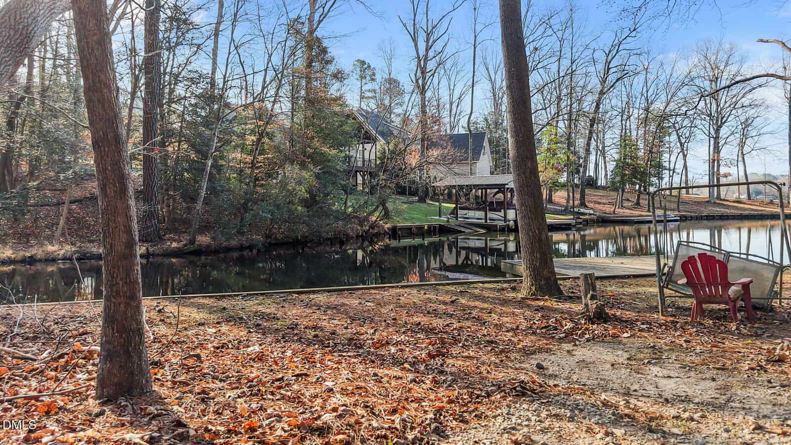 A wooded lakeside area with fallen leaves, a red Adirondack chair, a small dock, and a house in the background beside calm water, on a clear, sunny day.