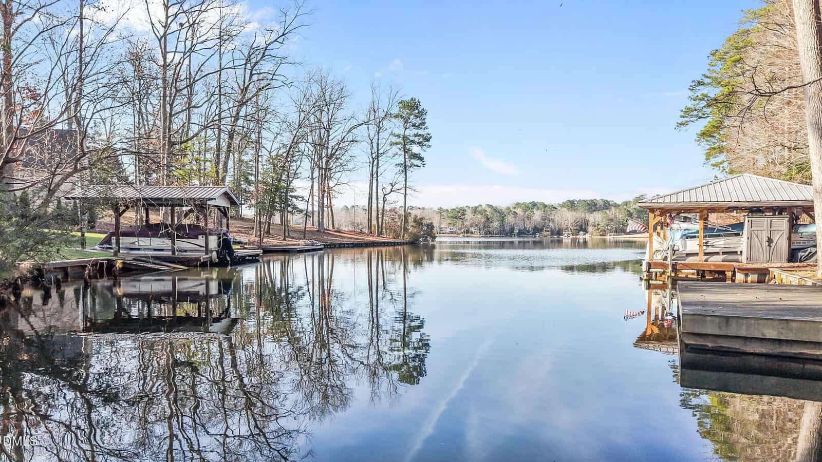 A calm lake bordered by trees, with two covered boat docks on either side at 1175 Sagamore Drive, Louisburg. Boats are moored under the docks, and bare branches reflect on the water. The sky is clear with a few clouds, signaling a peaceful day.
