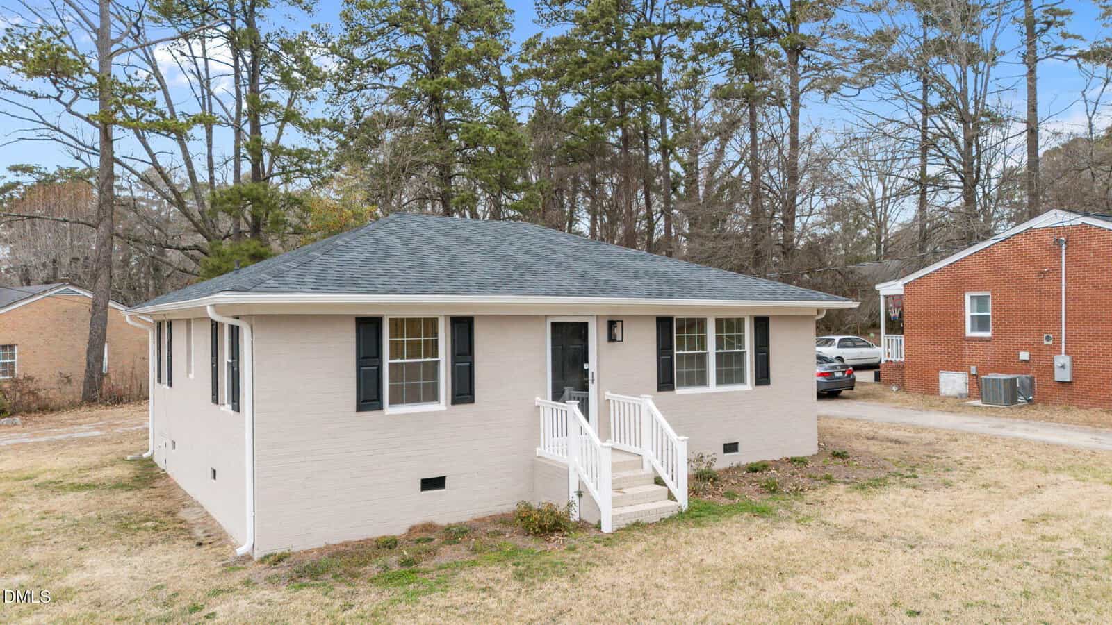 Single-story beige brick house with a dark shingle roof, white trim, small front porch, black shutters, and a car parked behind. The yard has patchy grass and is surrounded by trees and neighboring houses.