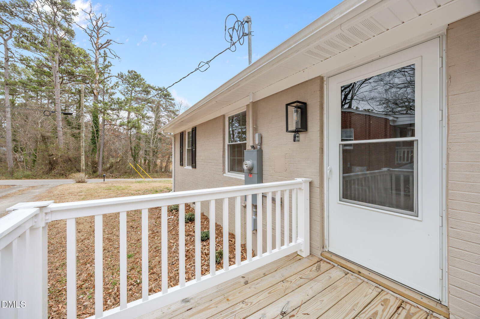 A beige brick house with a white framed storm door, white railing, and small porch. There are brown leaves on the ground and tall trees in the background under a blue sky.