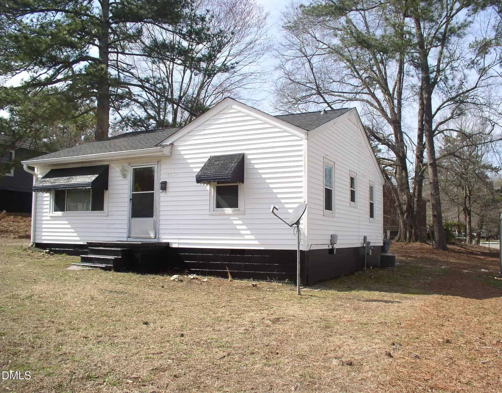 A small, single-story white house with black trim, black awnings over the windows, and a black foundation sits at 1061 N Pinkston Street in Henderson. The yard is mostly bare with a few trees, and there is a satellite dish near the house.