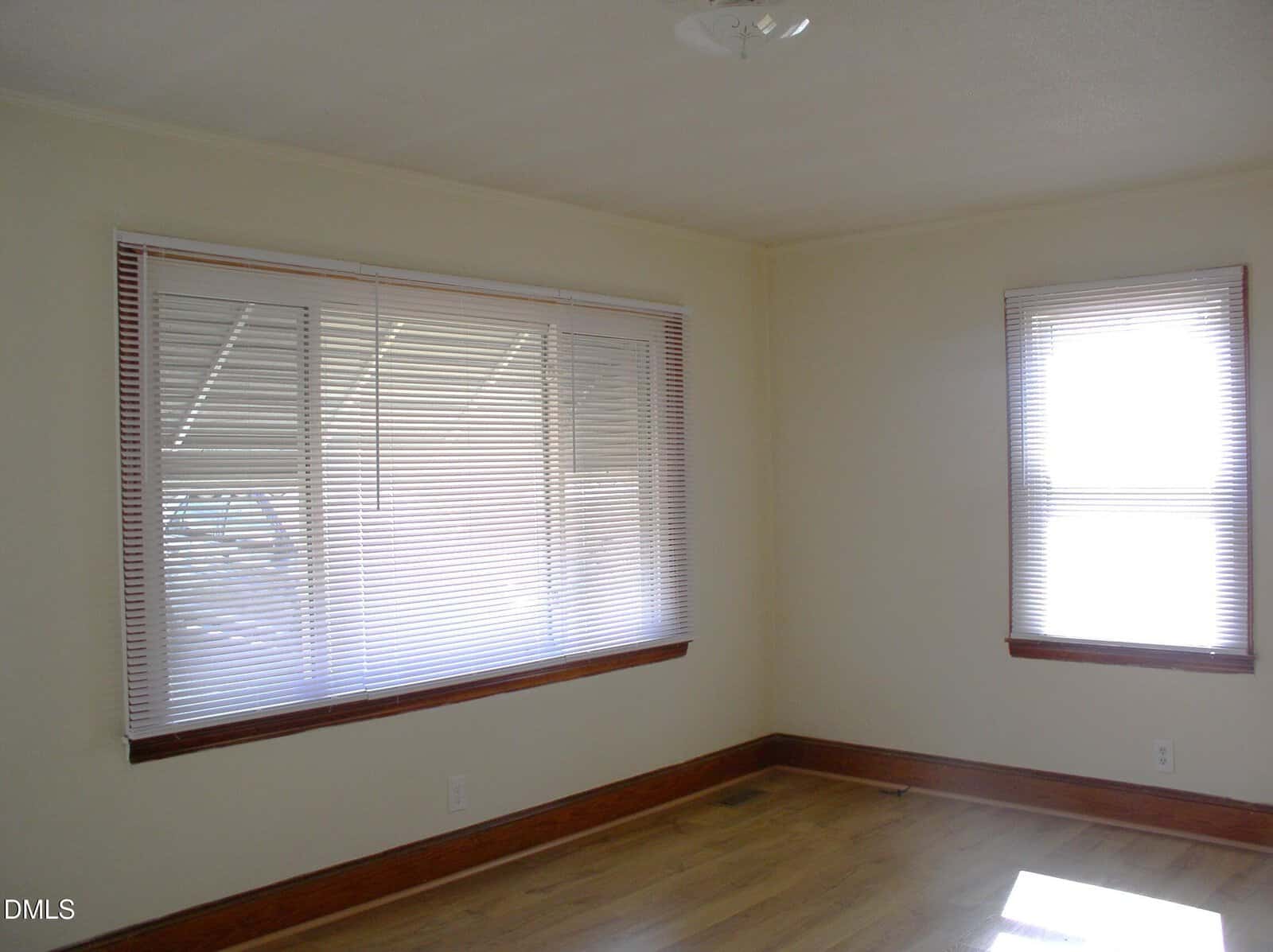 An empty room with light-colored walls, two windows with closed blinds, wooden trim, and a hardwood floor. Sunlight enters through the blinds, casting light patterns on the floor.