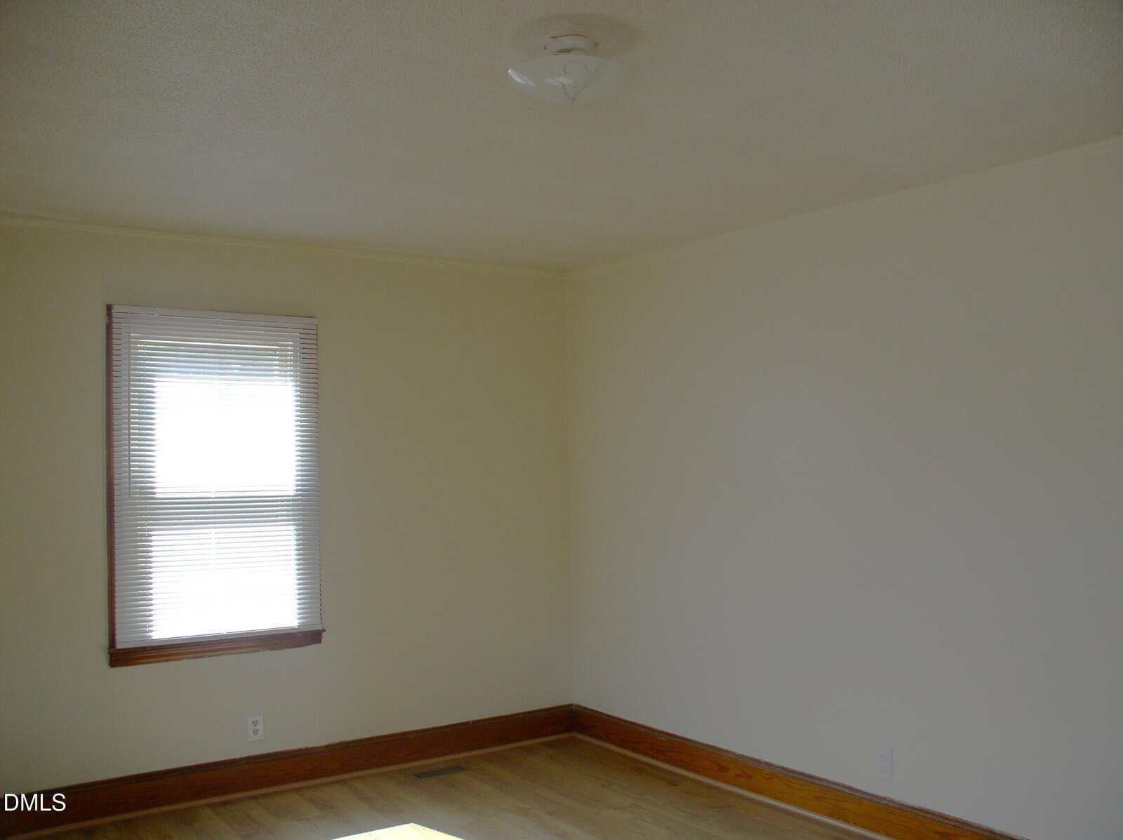 Empty room with cream-colored walls, a window with closed blinds, wood trim, a ceiling light fixture, and hardwood floors. Sunlight is coming through the window. The space appears clean and unfurnished.