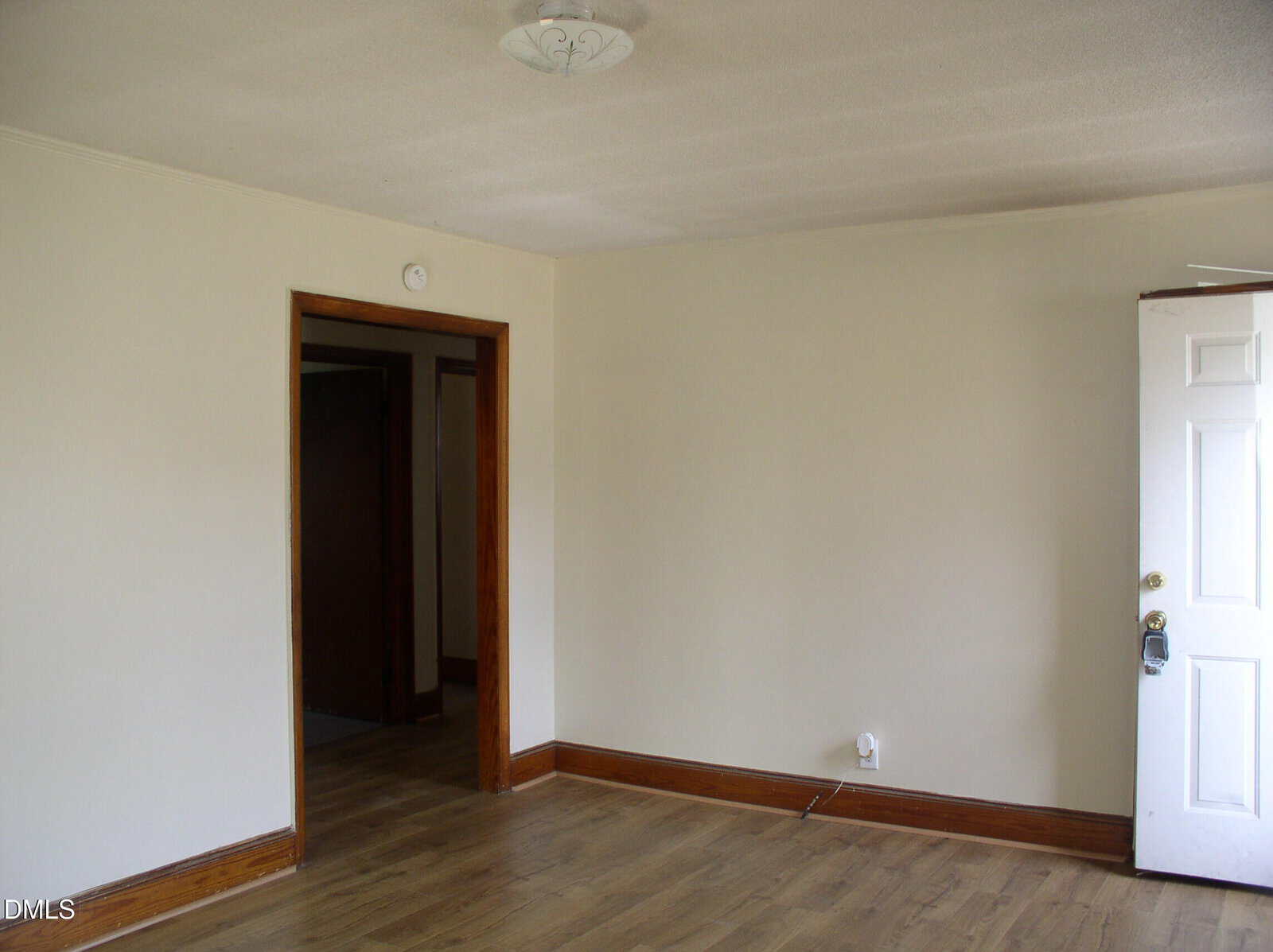Empty room with light beige walls, wood trim, and wood flooring. An open white door is on the right, leading outside. A doorway on the left opens to a hall with similar flooring and darker wood doors. Ceiling light fixture visible.