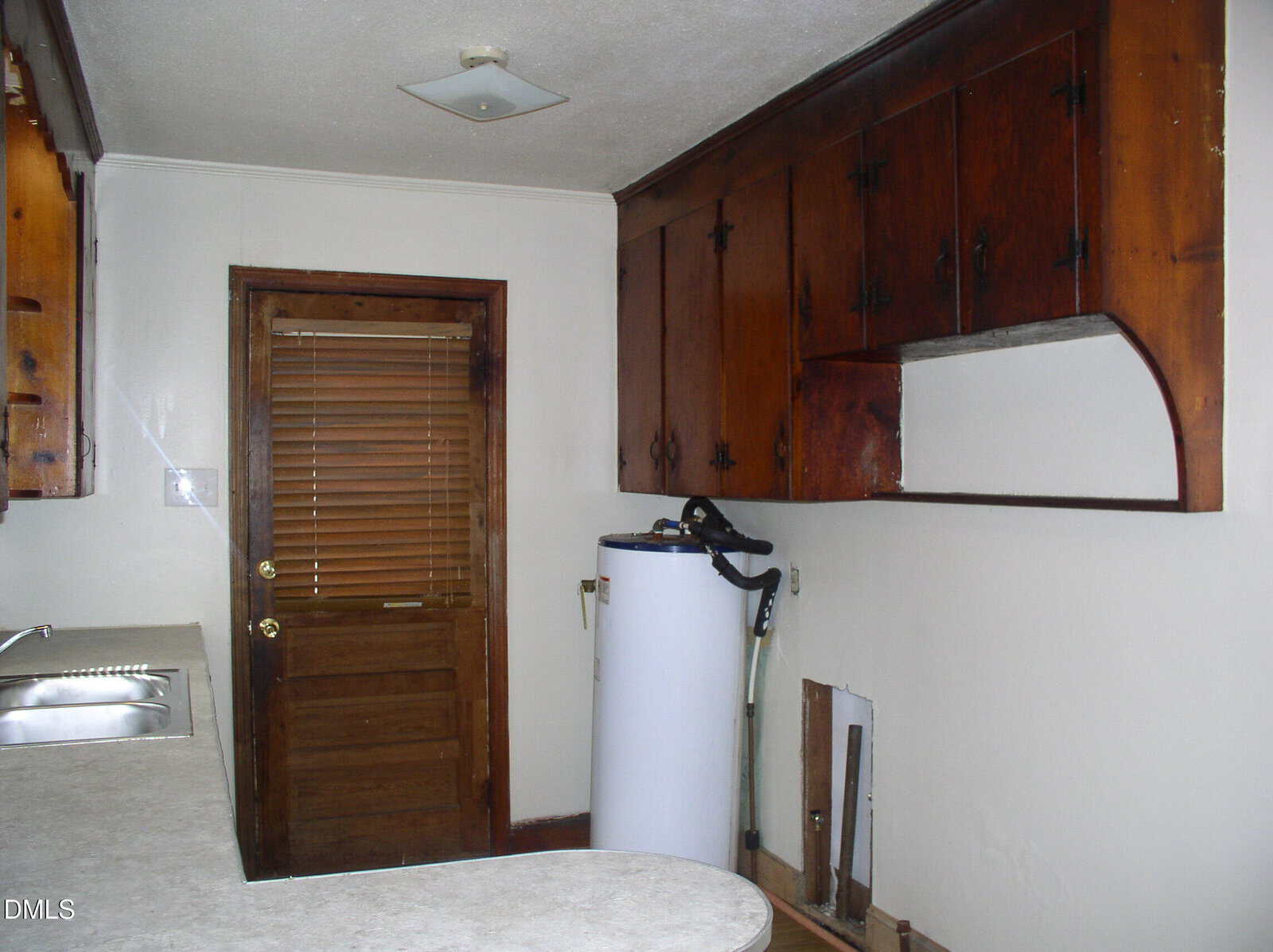 A small kitchen with wood cabinets, a stainless steel sink, a countertop, a water heater in the corner, a wooden door with blinds, and an unfinished section beneath the counter with exposed pipes.