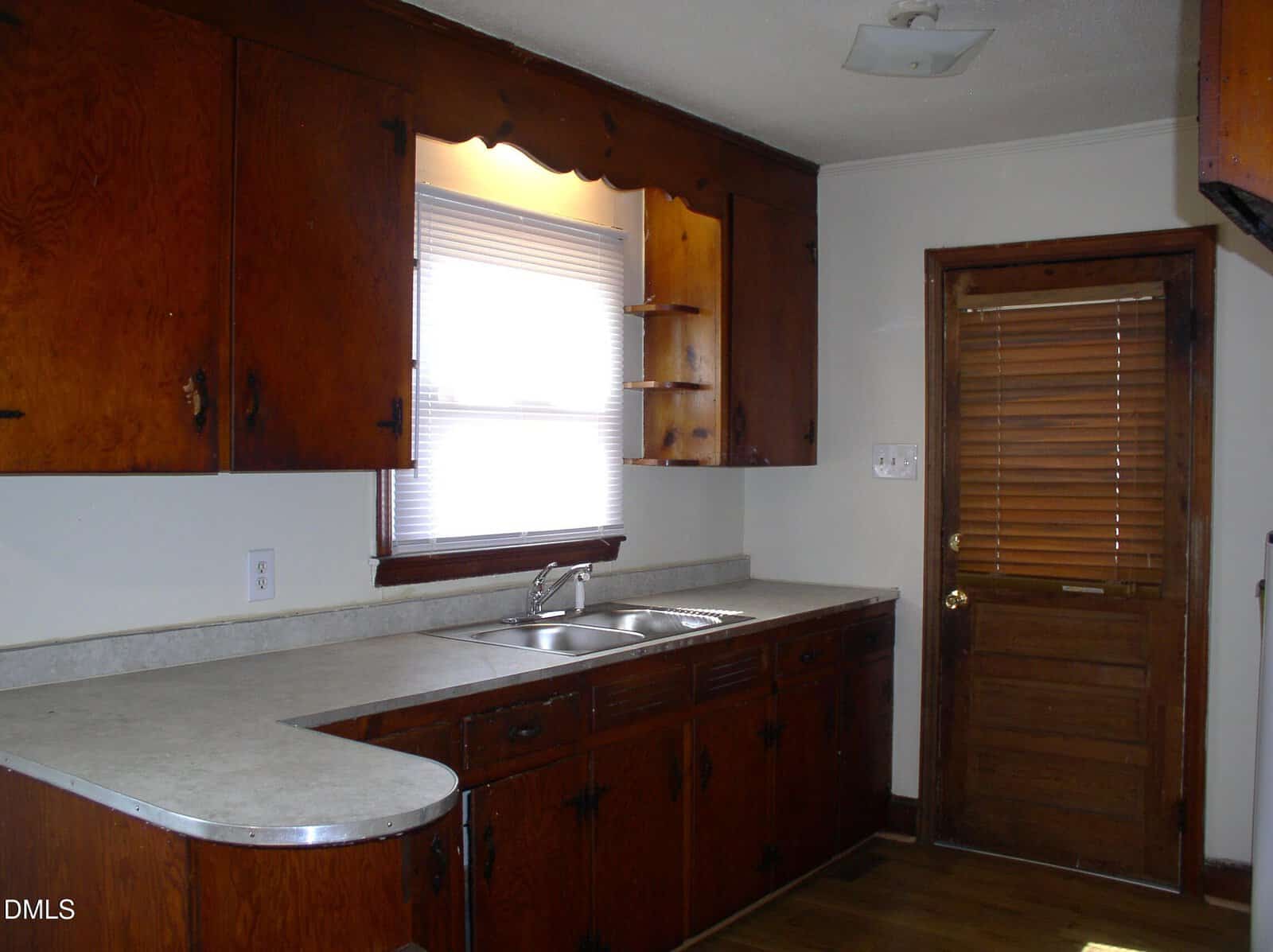 Kitchen with wood cabinets, a window above a double sink, white countertop, and a wooden door with blinds. There is no visible decor or appliances, and the space is lit by natural light.
