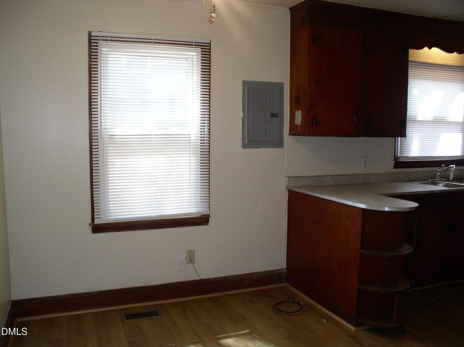 A window with closed blinds is next to a gray electrical panel on a cream wall. Wooden cabinets, a counter, and a sink are partially visible on the right. The floor is wood.