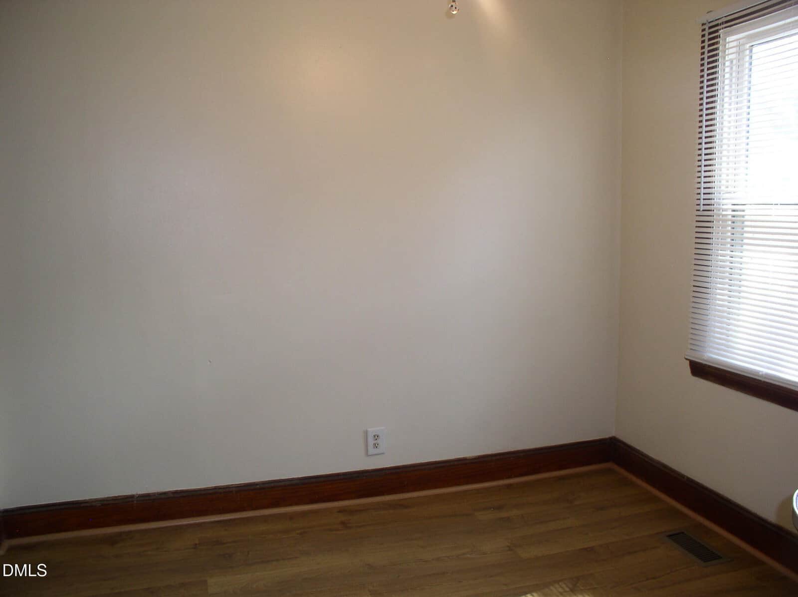 Empty room with beige walls, hardwood floor, a single electrical outlet, wood trim, and a window on the right with blinds partially open letting in natural light.