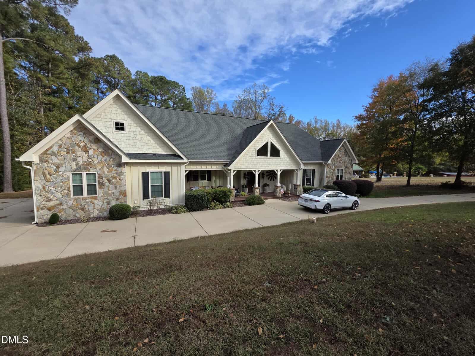 A single-story house with stone and light-colored siding, a covered front porch, and a gray roof sits beside a wide driveway with a white car parked in front. Trees with autumn foliage surround the property.