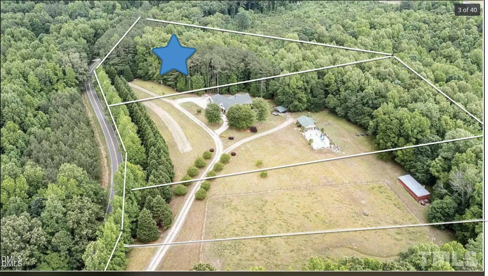 Aerial view of a wooded rural property outlined in white, featuring a house, a driveway, a smaller building, and a red shed, surrounded by trees and open grassy areas. A large blue star marks a section near the house.