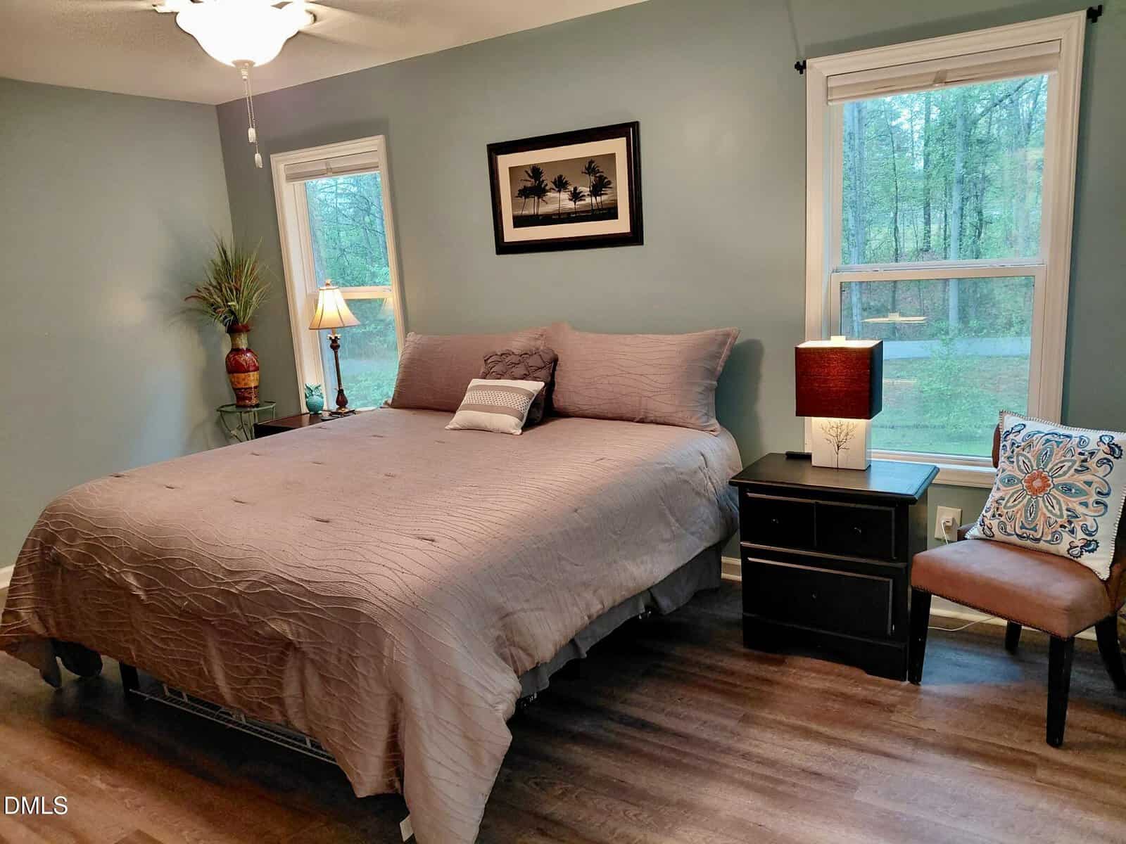 A bedroom with a neatly made bed, two windows, a nightstand with lamps, a plant, a chair with a decorative pillow, and wall art above the bed. The room has wood flooring and soft blue walls.