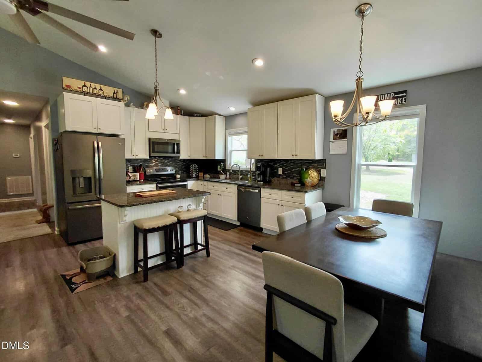 Modern kitchen with white cabinets, stainless steel appliances, a central island with two stools, and a dining table with four chairs. Large window lets in natural light; wood-look flooring throughout.