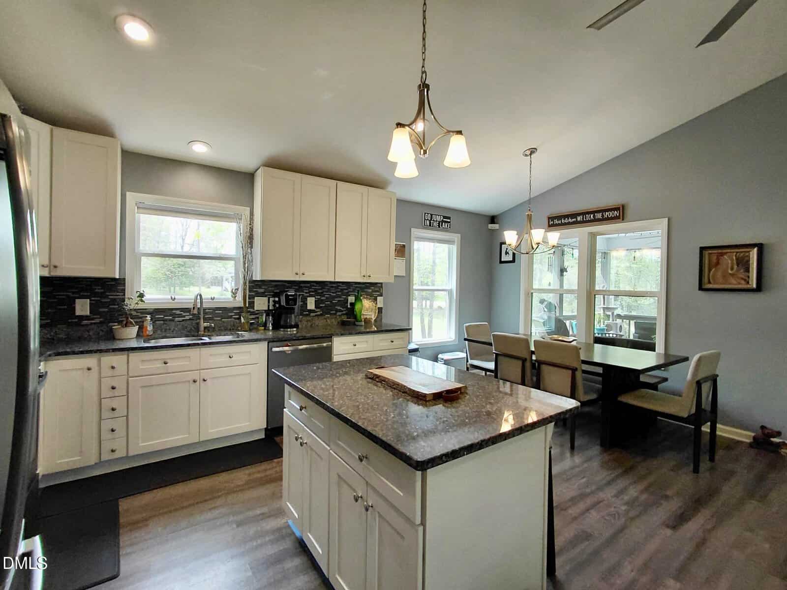 Modern kitchen and dining area with white cabinets, a central island with dark countertop, stainless steel appliances, large windows bringing in natural light, and a dining table with six chairs near a glass patio door.