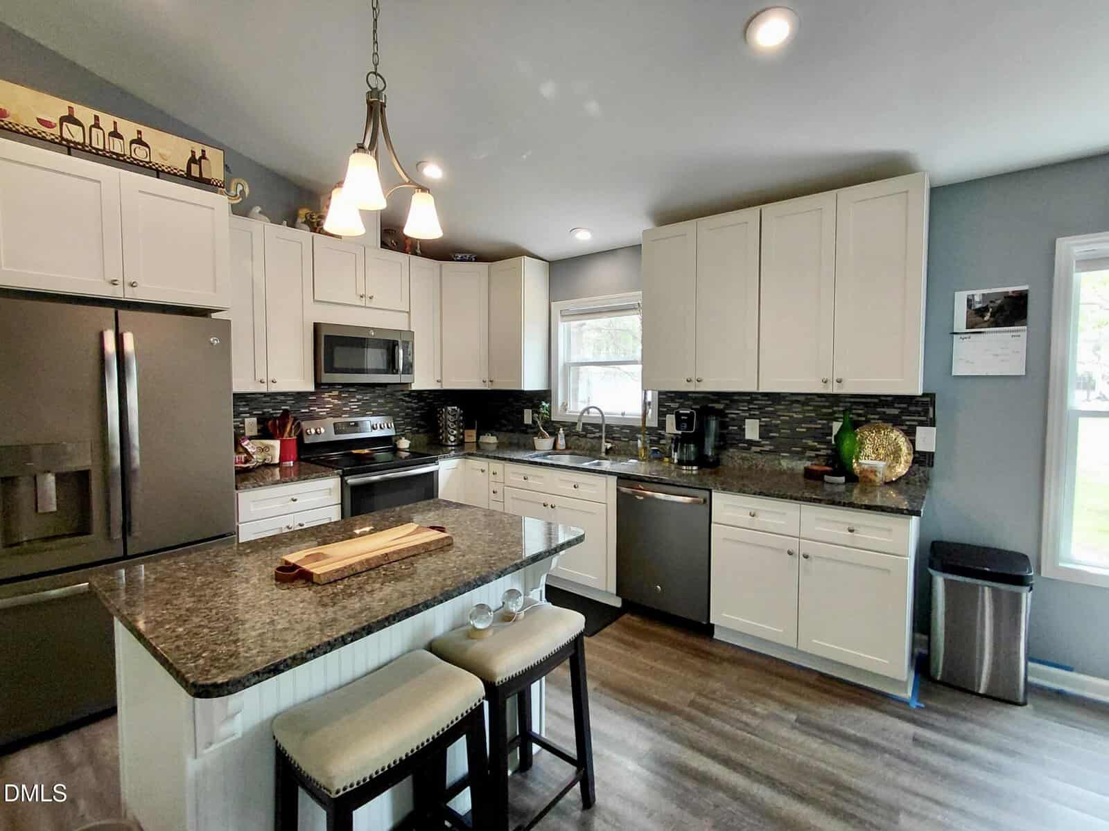 Modern kitchen with white cabinets, stainless steel appliances, dark countertops, an island with two stools, pendant lights, and a window letting in natural light. Decorative items and plants accent the space.