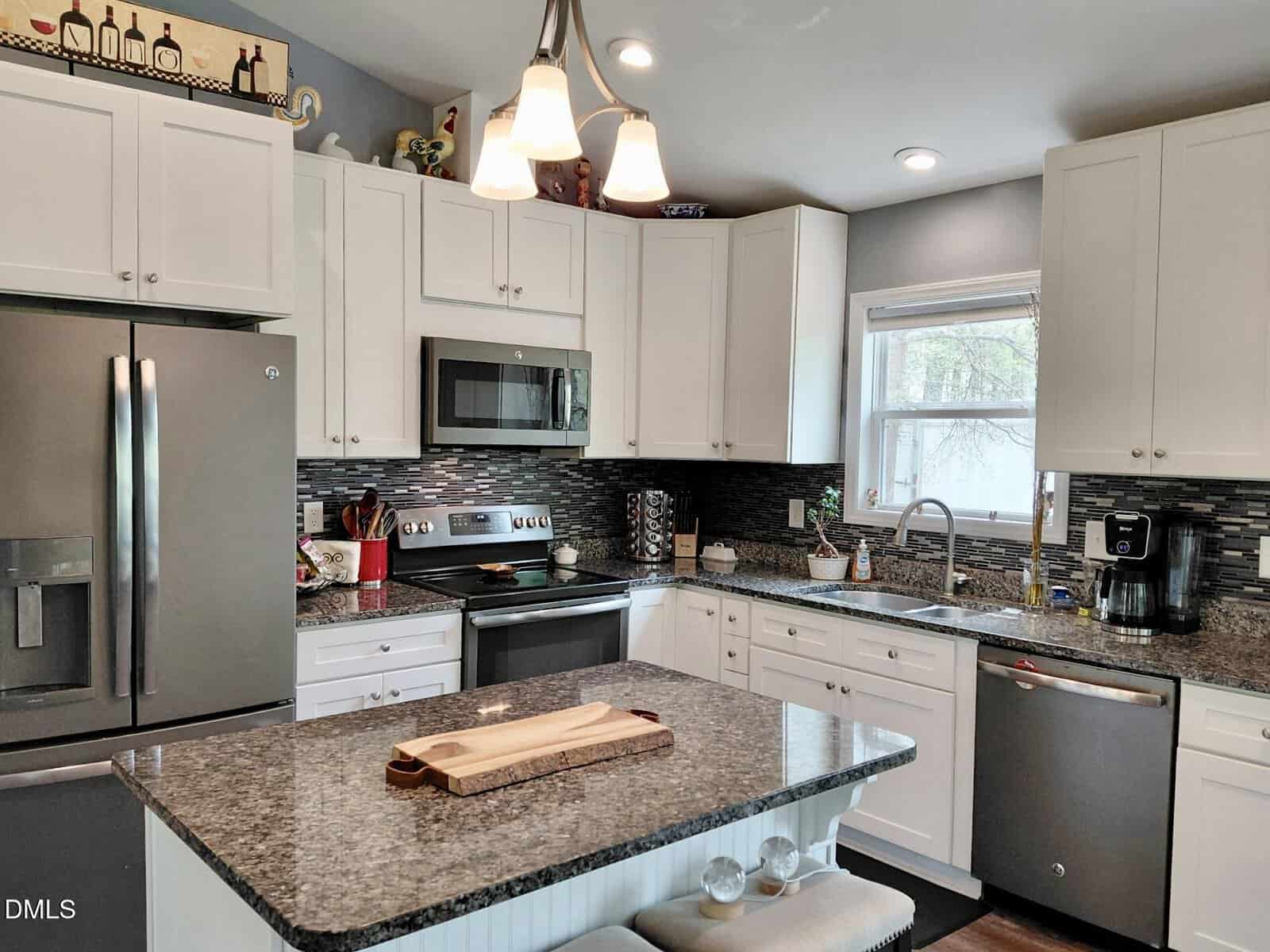 Modern kitchen with white cabinets, stainless steel appliances, granite countertops, a center island with a wooden tray, and black tile backsplash. Pendant lights hang above the island. Window over the sink lets in natural light.