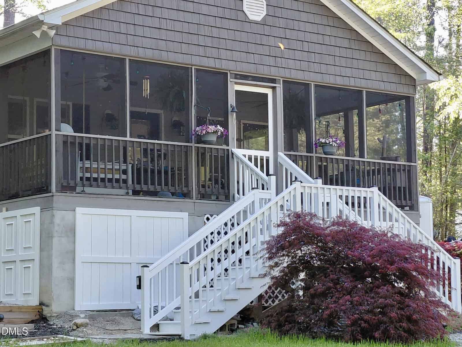 A screened-in porch with dark railings and hanging flower baskets sits above a white staircase, next to a red-leafed bush and surrounded by trees. The area beneath the porch has white paneling and a door.