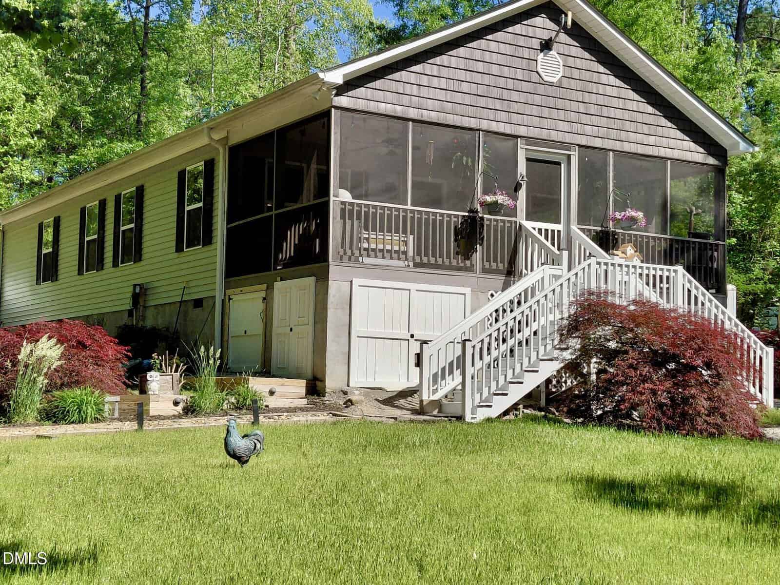 A single-story house with a screened porch, white railings, and stairs sits amid green trees and shrubs. A black chicken stands on the front lawn near red-leafed bushes.