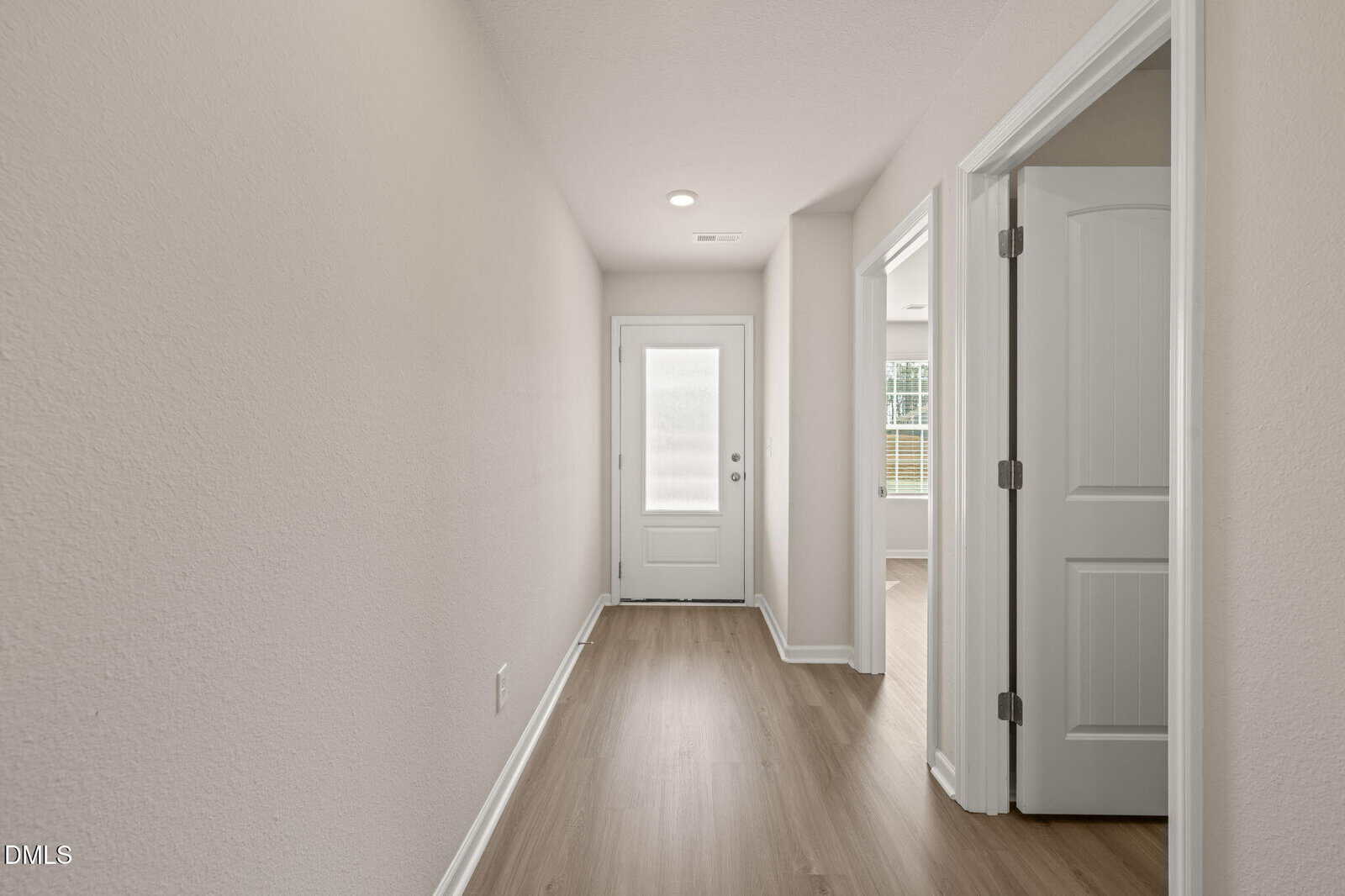 A hallway with light wood flooring, light beige walls, and white trim leads to a closed white door with a frosted glass panel. Two open doors on the right reveal rooms with similar flooring and natural light.