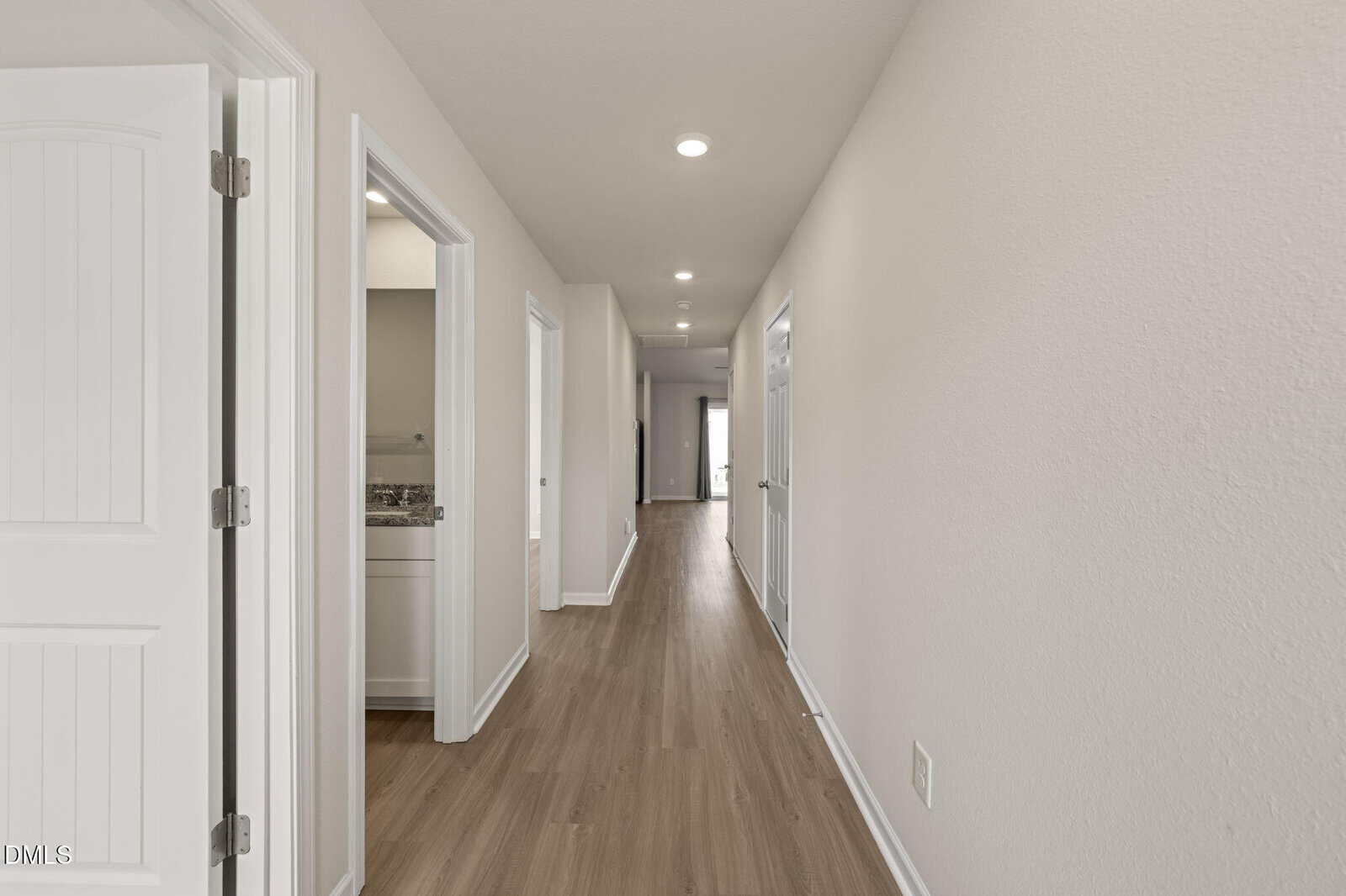 A long, empty hallway with light-colored walls, wood flooring, ceiling lights, and white doors on both sides. At the end of the hallway, natural light is visible from a distant window or glass door.