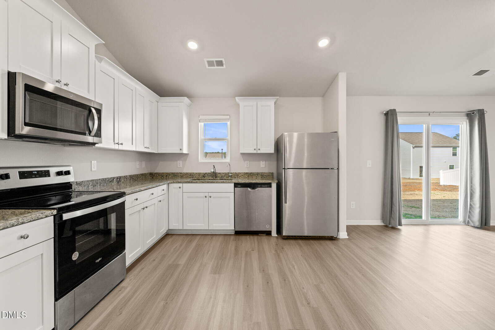 Modern kitchen with white cabinets, granite countertops, stainless steel appliances, light wood flooring, a small window above the sink, and glass doors leading to a backyard. The space is bright and unfurnished.